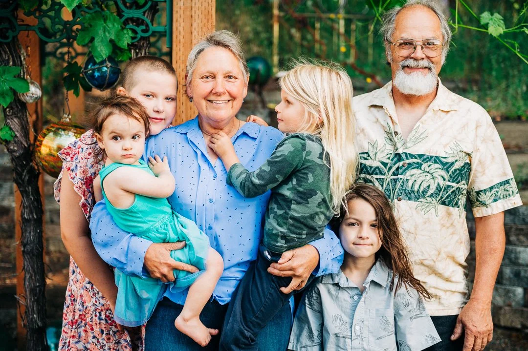 A multigenerational family of grandparents with five grandchildren standing together outdoors in a garden, smiling at the camera.