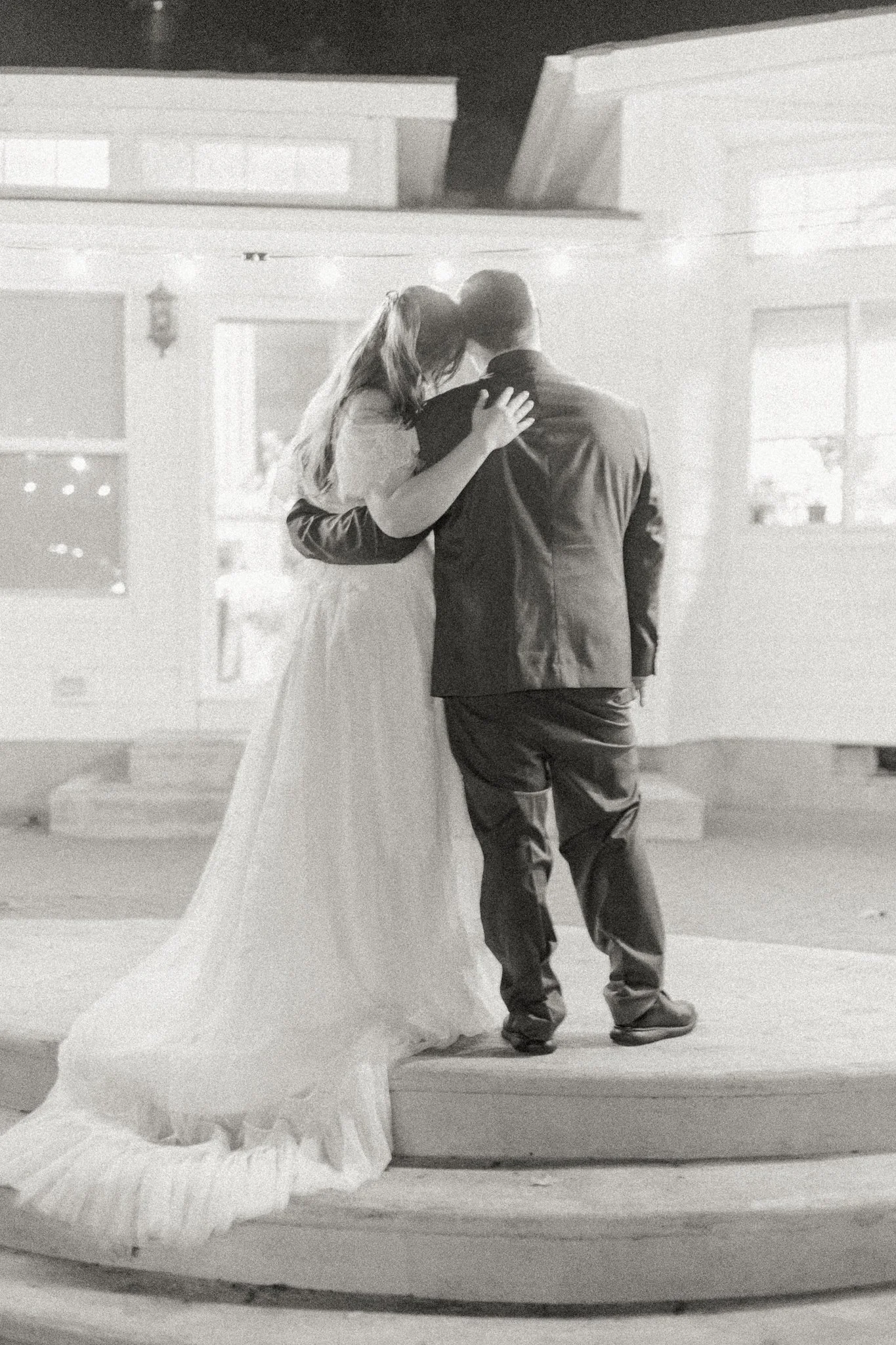 A black and white photo of a bride and groom sharing a kiss, with the bride in a wedding dress and the groom in a suit, standing on a small stage.