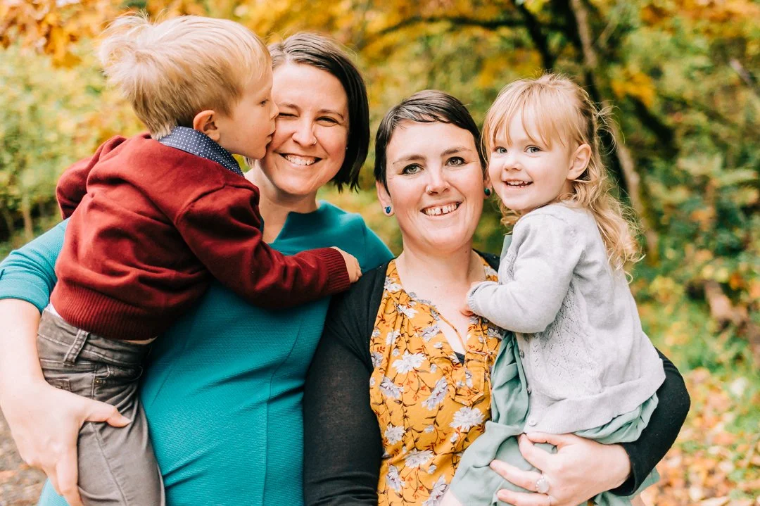 Two women holding children outdoors during fall, smiling at the camera, with autumn trees and foliage in the background.