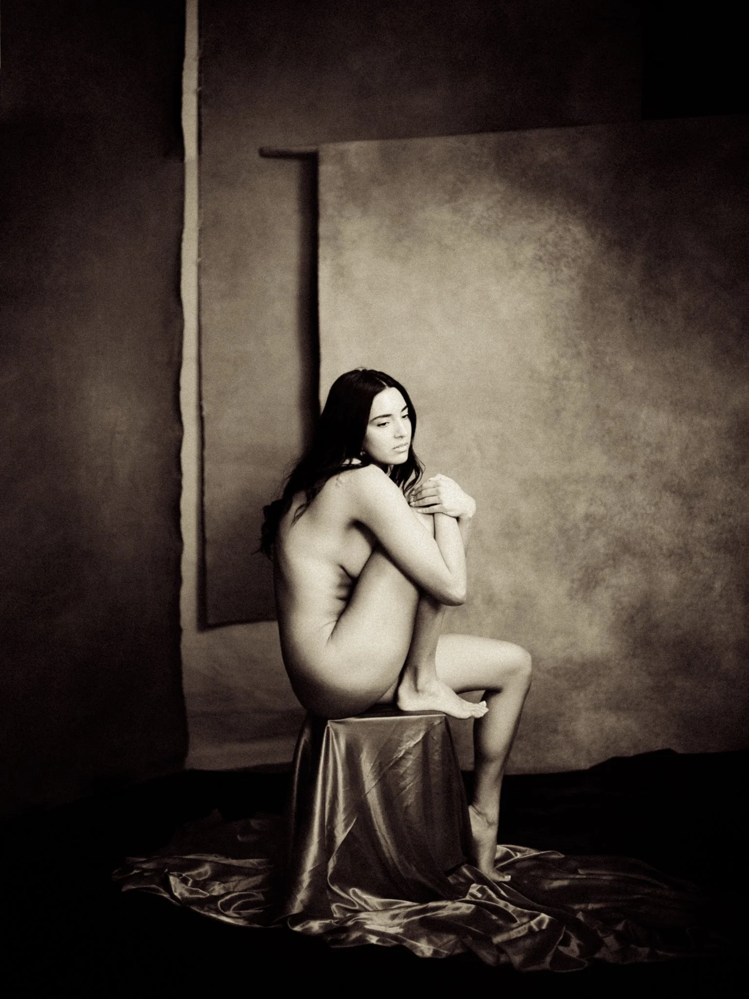 Black and white photo of a nude woman sitting on a stool, hugging her knees, with a personified textured backdrop behind her.