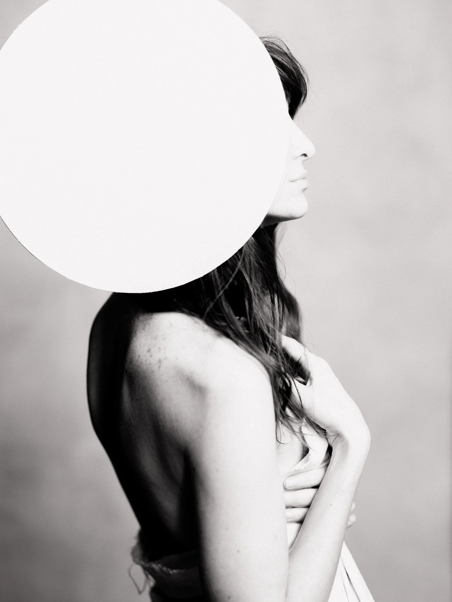 Black and white photo of a woman with long hair, holding her chest, with part of her face covered by a large white circle.