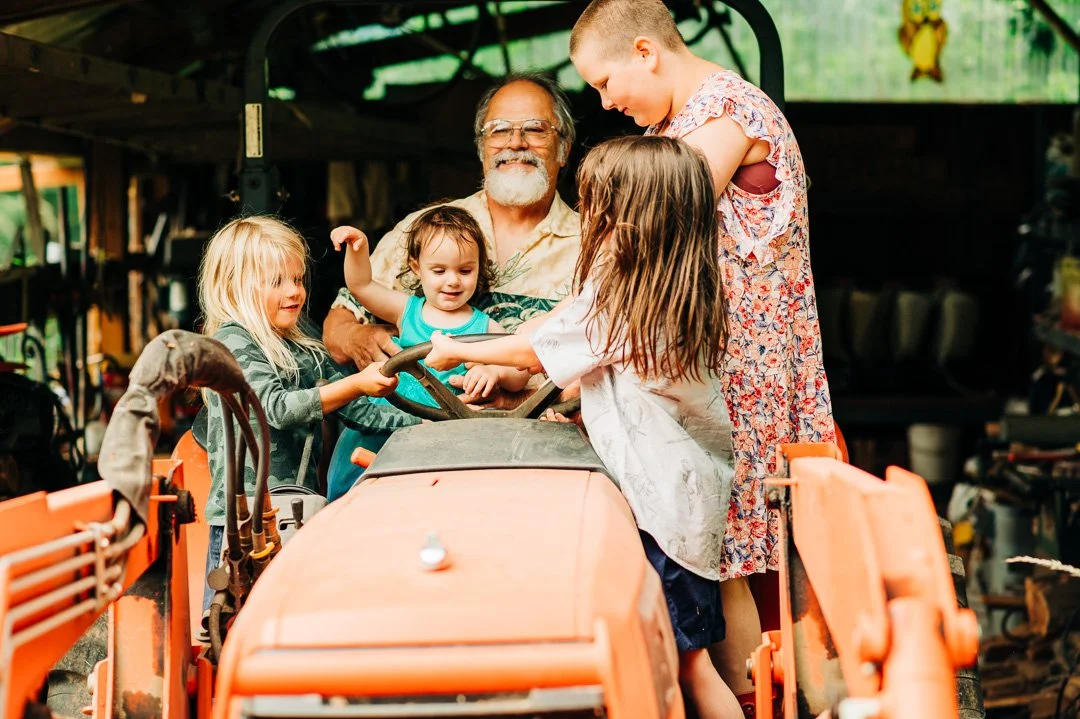 Children and an elderly man on a tractor in a garage or workshop.