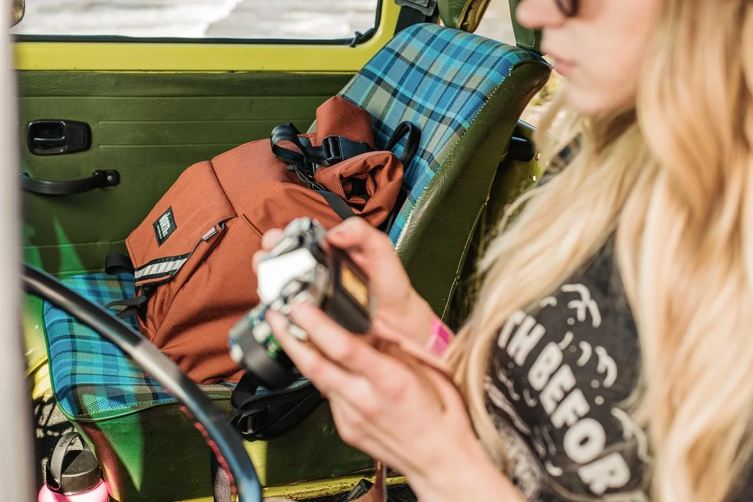 A woman with long blonde hair looking at a camera inside a vehicle. There is an orange backpack on the green plaid seat next to her, and part of a pink water bottle is visible in the lower left corner.