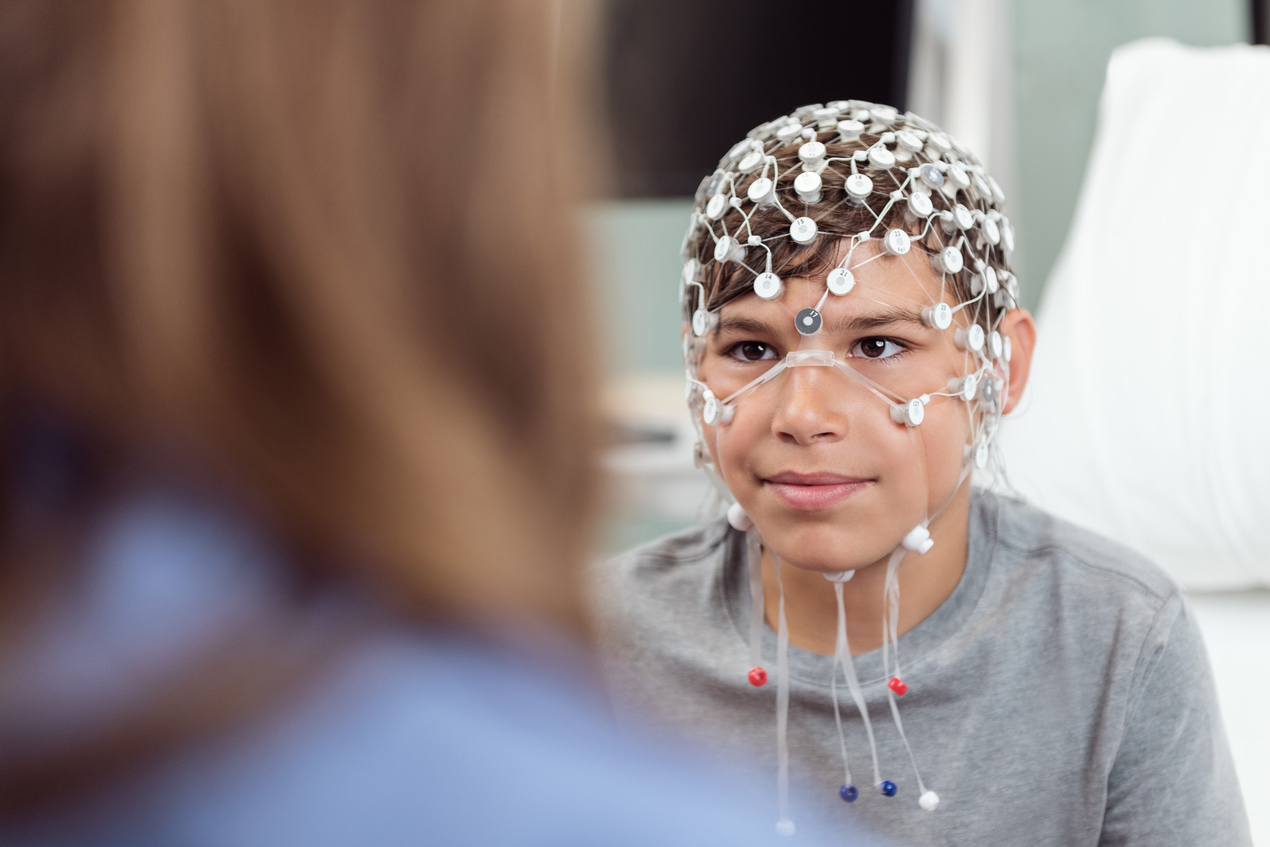 Child in a hospital wearing an electroencephalogram (EEG) cap with numerous electrodes, with a caregiver partially visible in the foreground.