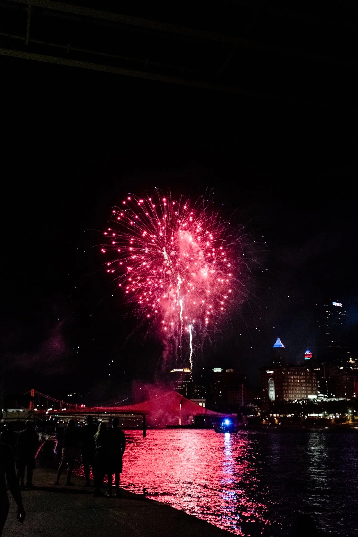 Fireworks exploding over a city skyline at night, with reflections on the water and silhouetted people watching from the waterfront.