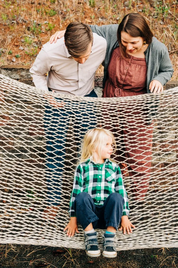 Two adults assisting a child sitting on a rope hammock outdoors.