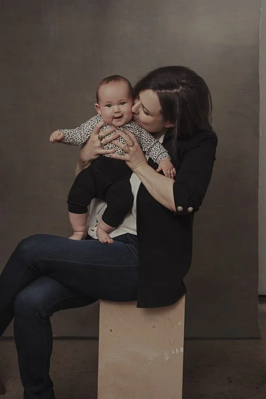 A woman sitting on a wooden block holding a happy baby with a patterned shirt and black pants, against a plain brown background.