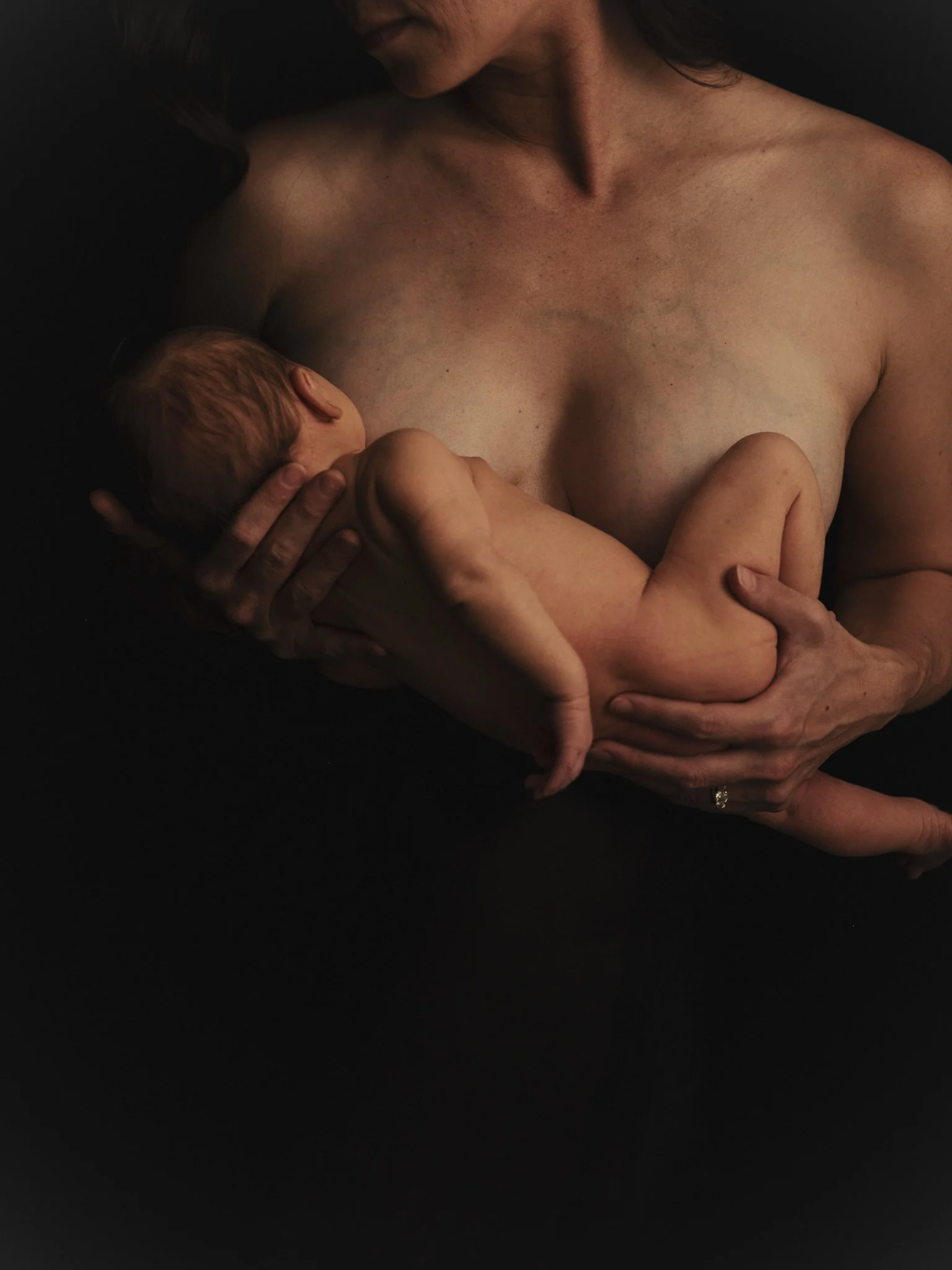 A woman breastfeeding a newborn baby against a dark background.