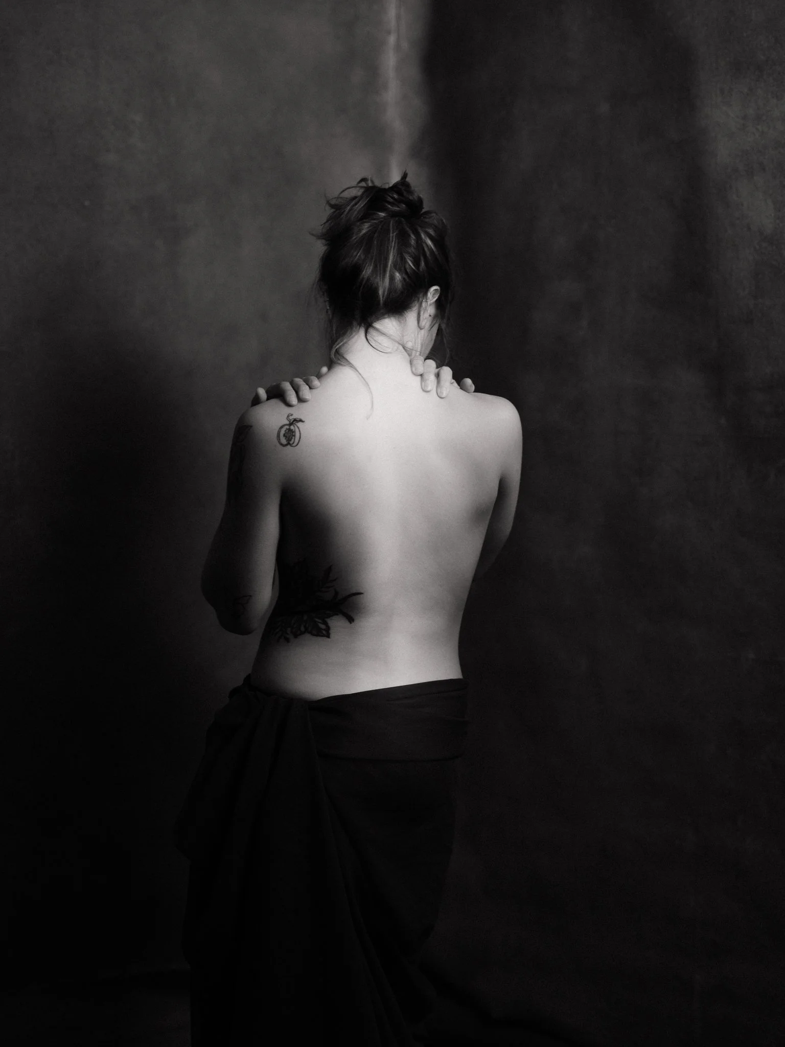 Black and white photo of a woman seen from behind, topless with tattoos on her left shoulder and side, wearing a dark cloth wrapped around her waist, with her hands on her neck.