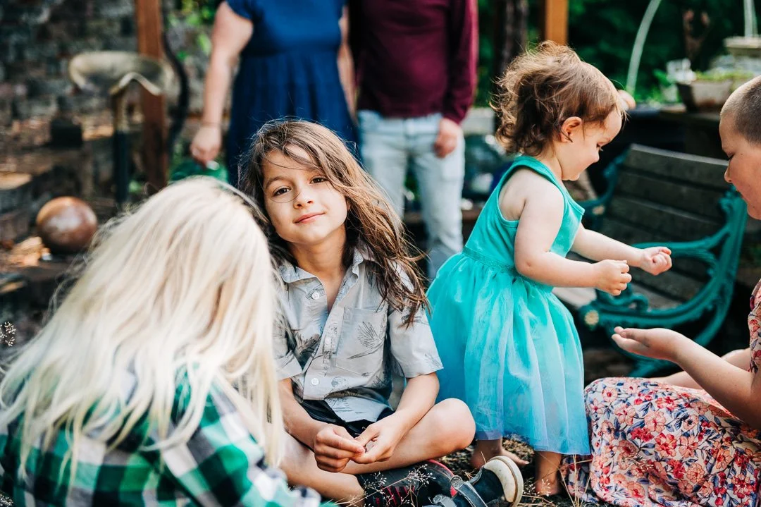Children sitting and playing outdoors, with adults in the background, in a garden or backyard setting.