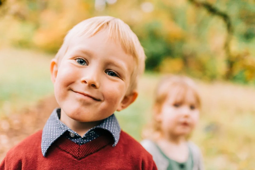 A young boy with blonde hair and blue eyes smiling outdoors during autumn, wearing a red sweater over a blue collared shirt, with a blurred girl in the background.