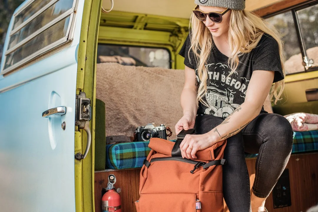 A young woman with blonde hair wearing sunglasses, a gray beanie, and a black T-shirt, sitting inside a vintage yellow and blue camper van, packing an orange backpack with a camera nearby.