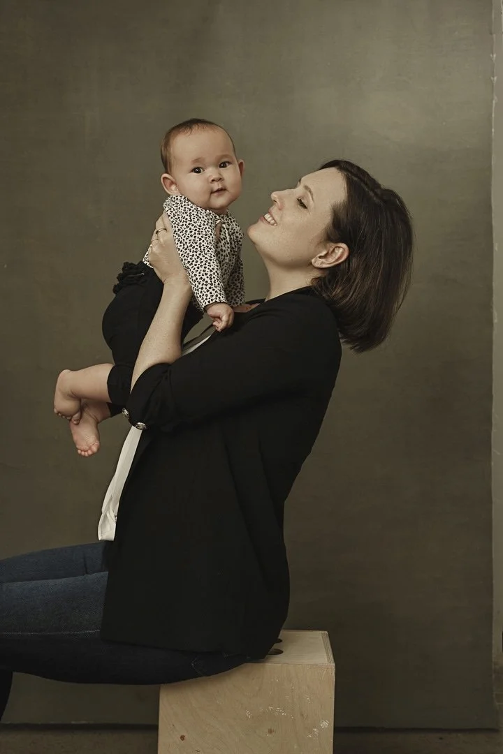 A woman with shoulder-length dark hair sitting on a wooden block, holding and looking lovingly at a baby girl with short hair and a blue and white polka-dot shirt against a plain brown backdrop.