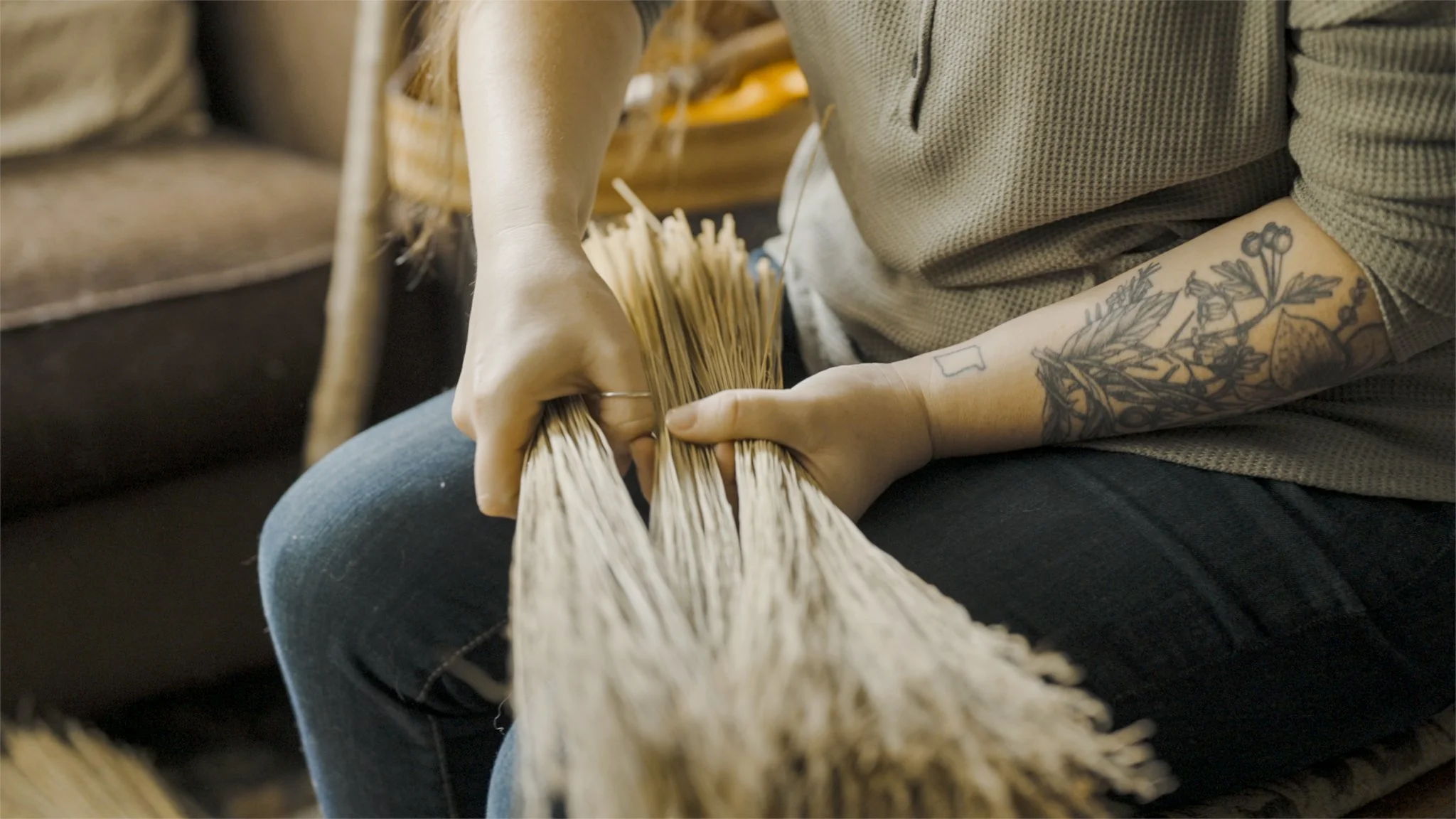 Person weaving straw, with tattooed arm, sitting on a chair.