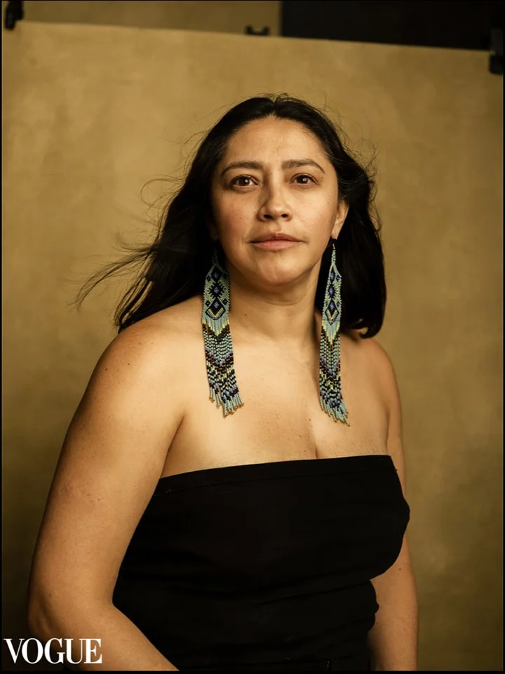 A woman with long dark hair wearing a strapless black top and large beaded earrings, standing against a neutral background.