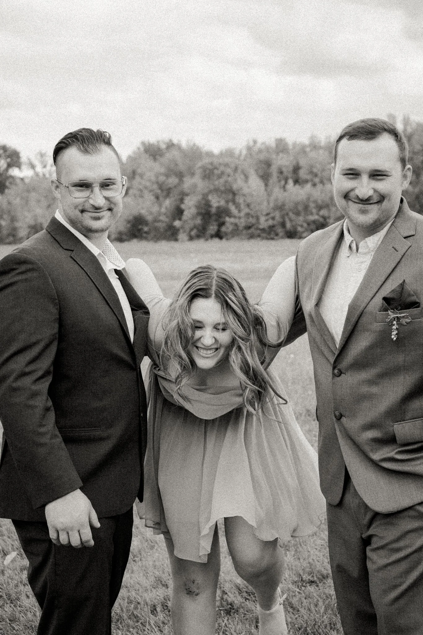 Three people smiling outdoors, two men in suits and a woman in a dress, with their arms around each other, in a natural setting with trees in the background.