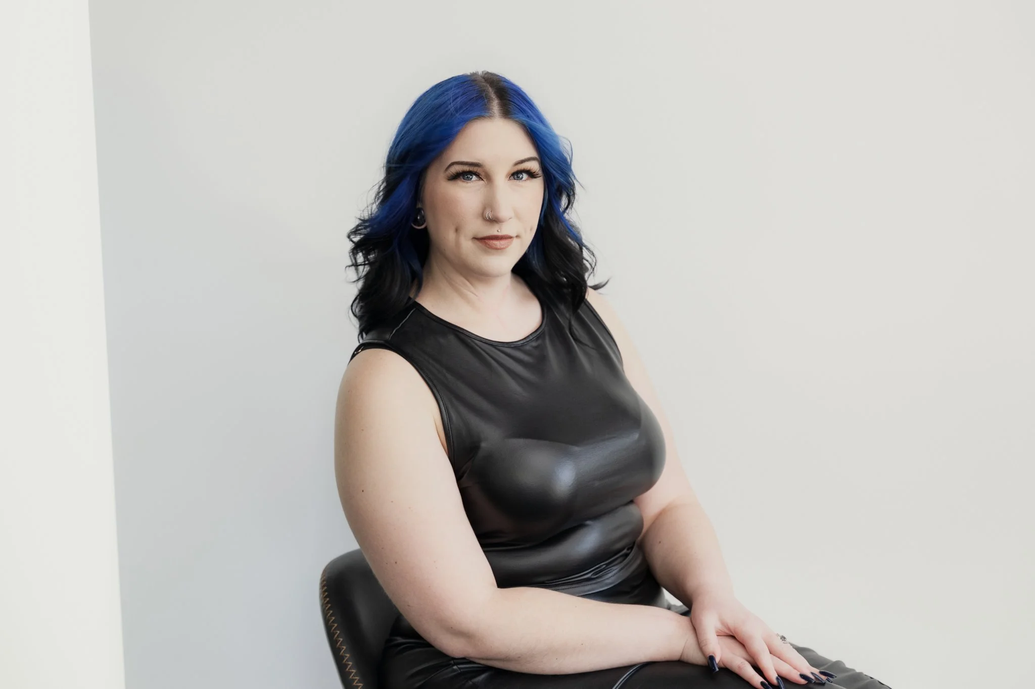 A woman with blue and black hair sitting on a black chair against a plain white wall, wearing a sleeveless black leather top.