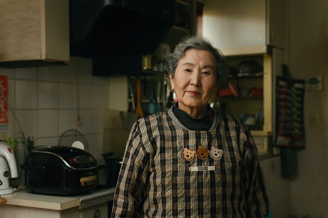 An elderly woman with gray hair standing in a kitchen, wearing a checkered apron with bear face buttons, looking at the camera.