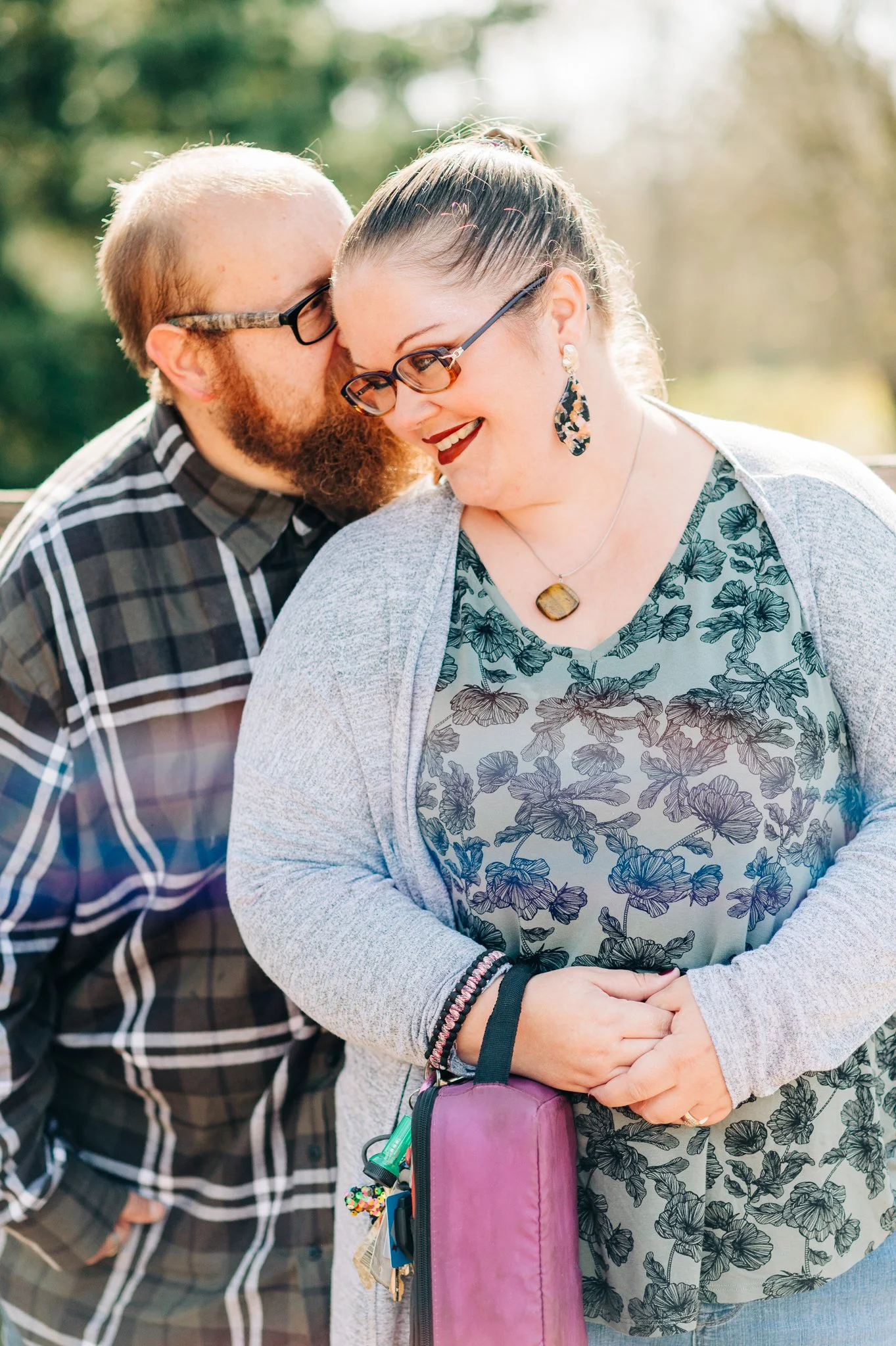 A man whispering into a woman's ear outdoors on a sunny day, both smiling. The woman is wearing glasses, a floral blouse, and earrings, and the man has a beard and glasses.