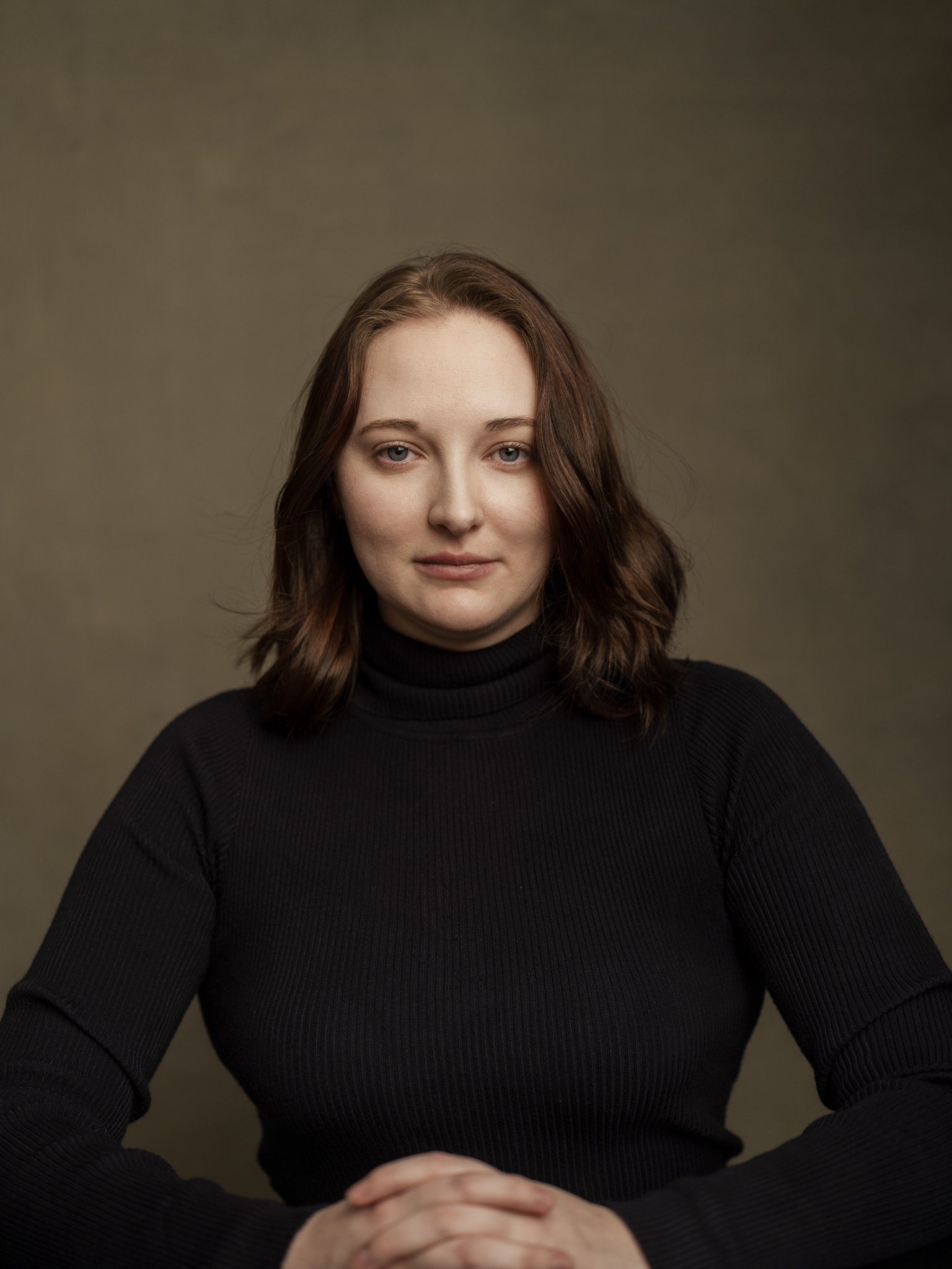 Portrait of a woman with shoulder-length brown hair wearing a black turtleneck, sitting with hands clasped in front of her against a plain muted background.