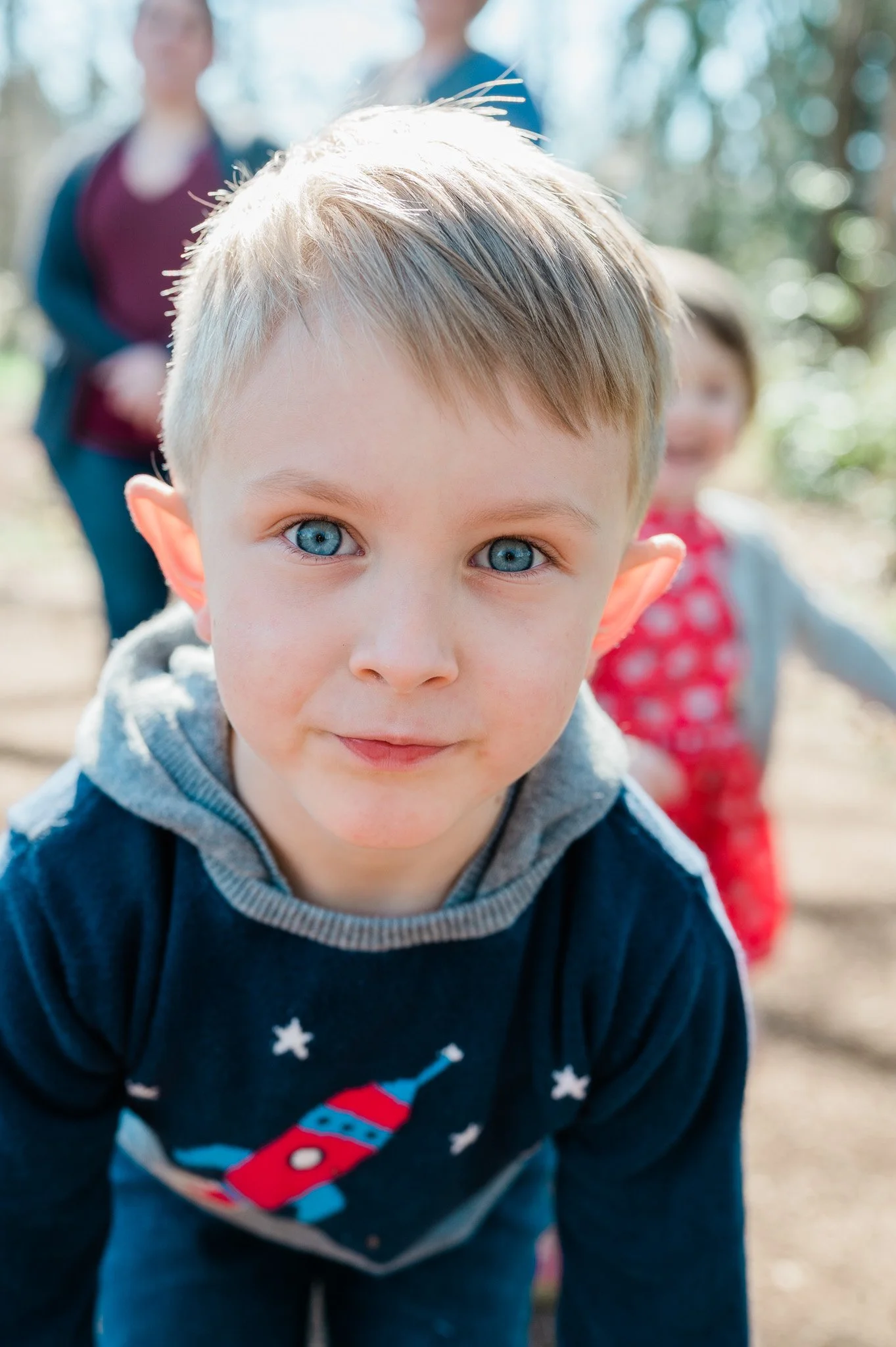 Close-up of a young boy with bright blue eyes and light brown hair, wearing a dark blue hoodie with a rocket ship design, outdoors with blurred trees and two other children and an adult in the background.