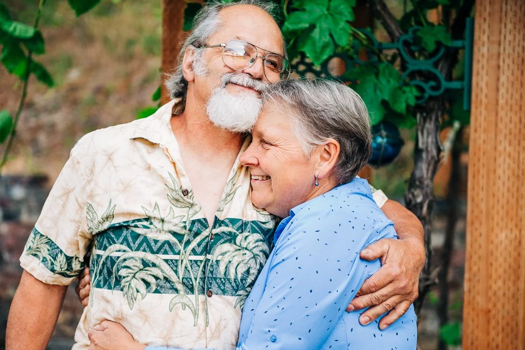 An elderly man and woman sharing a warm hug outdoors, smiling, with greenery in the background.