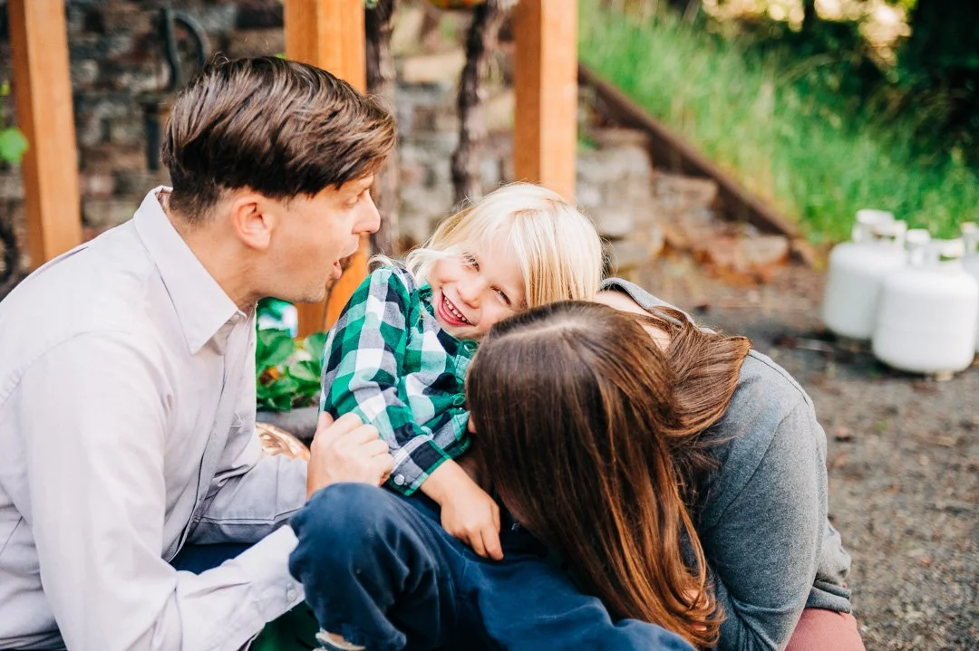 A family of three, consisting of a man, a woman, and a young girl, outdoors in a garden. The girl is laughing and playing, while the man and woman are engaging with her affectionately.