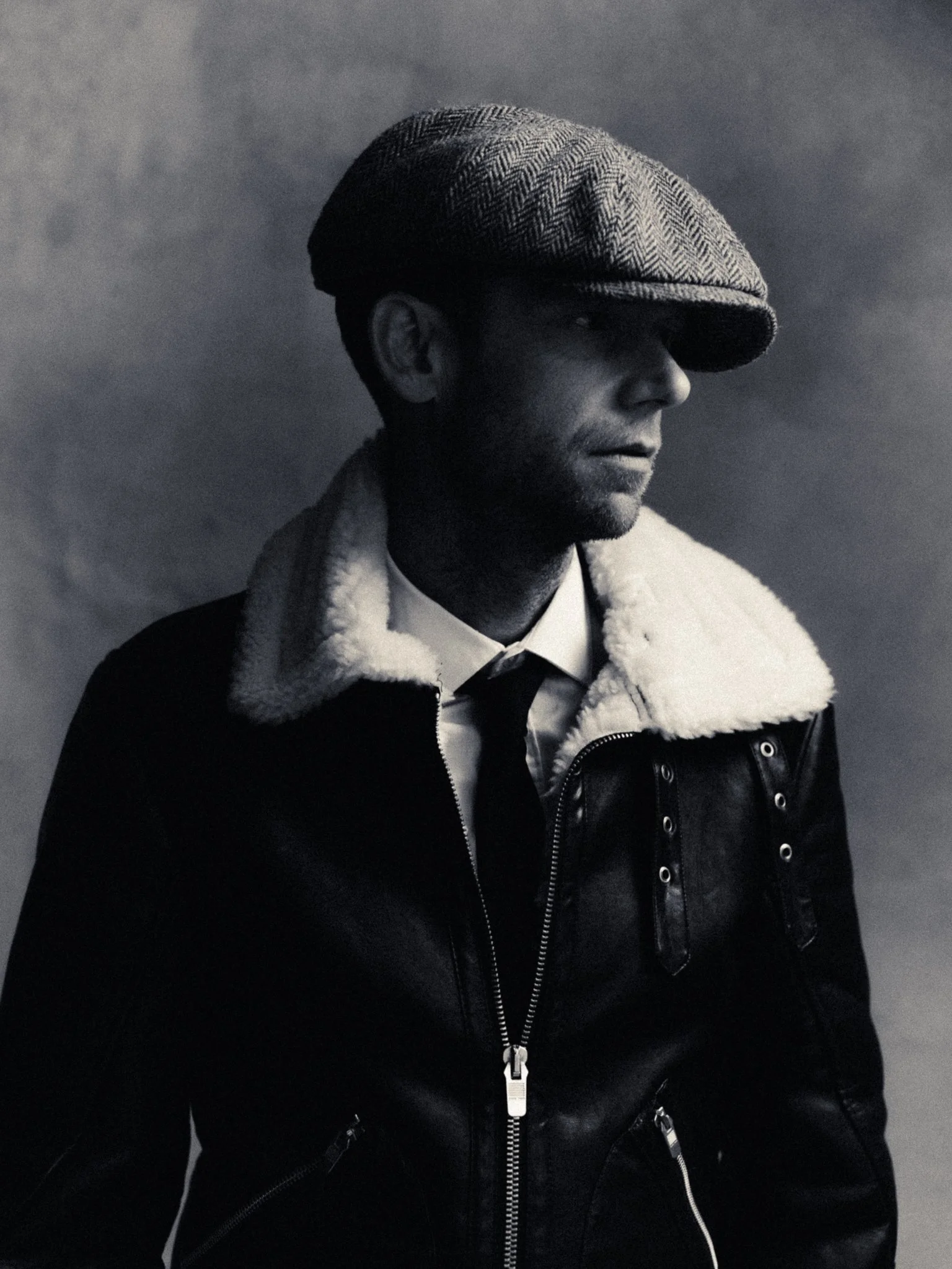 Black and white portrait of a man wearing a flat cap, a shearling-lined jacket, a shirt, and a tie, with a contemplative expression.