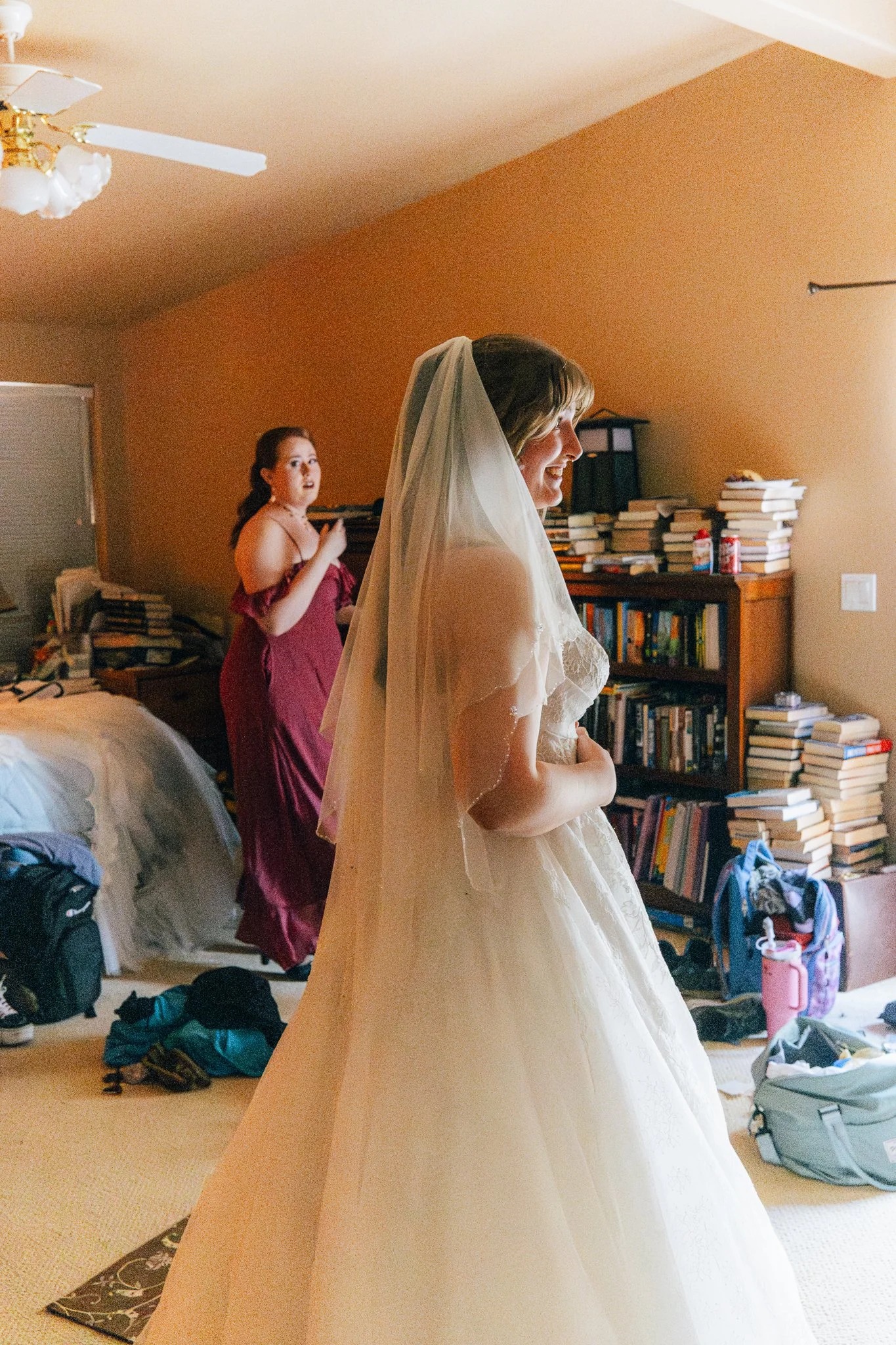 A bride in a wedding gown and veil smiling as she prepares for her wedding, with another woman in a burgundy dress behind her, in a cluttered room filled with books, bags, and clothes.