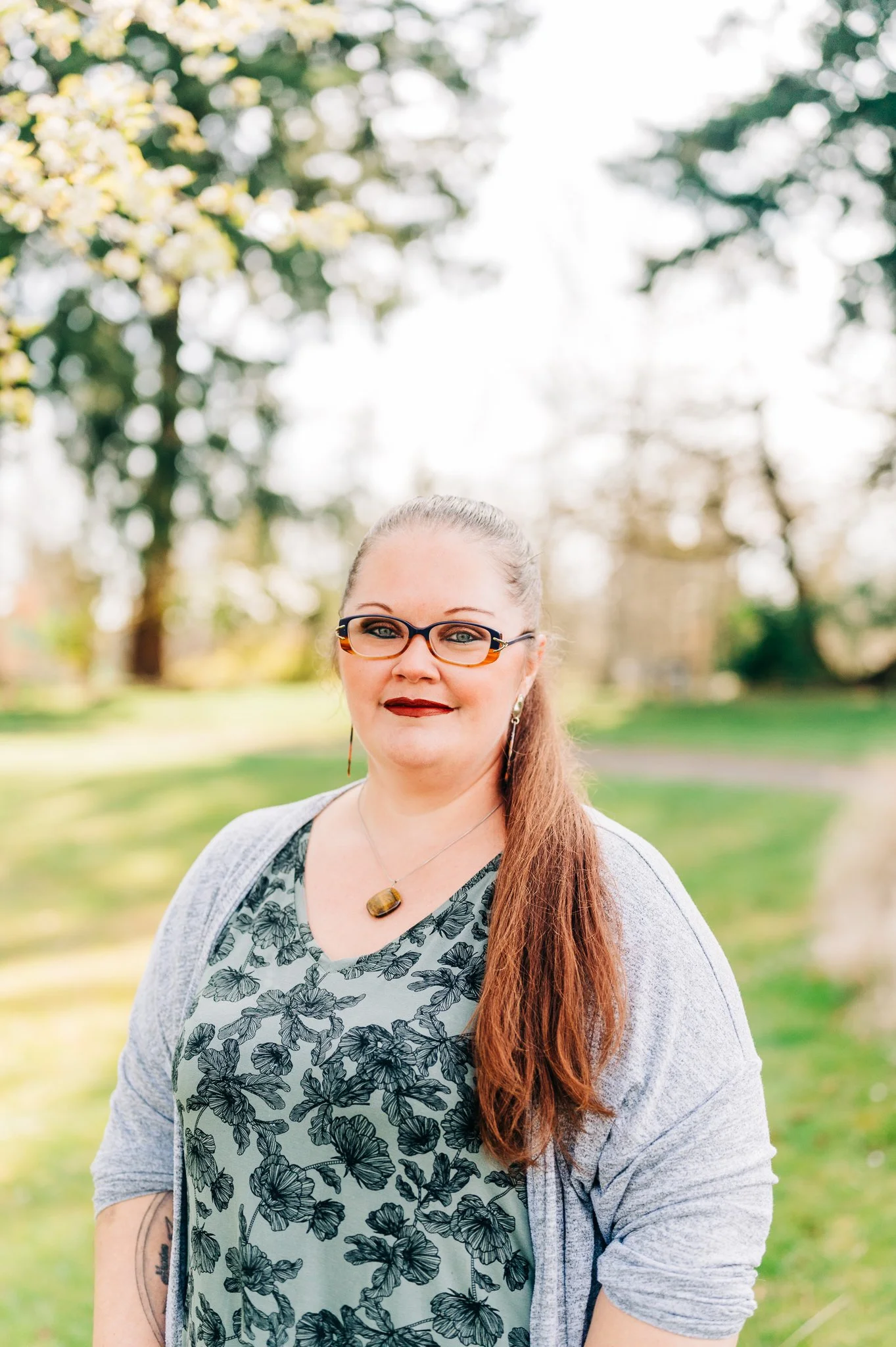 A woman outdoors wearing glasses, a floral-patterned shirt, a gray cardigan, and a pendant necklace, with long brown hair and earrings, standing in a park with trees in the background.