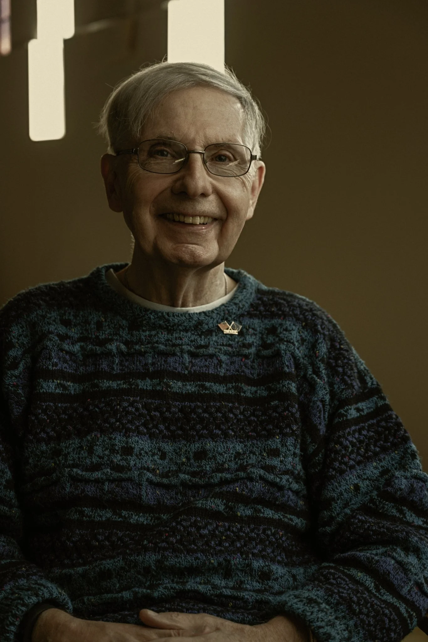 An elderly woman with short gray hair and glasses smiling, wearing a colorful patterned sweater with a small pin on her chest, sitting indoors with soft lighting in the background.