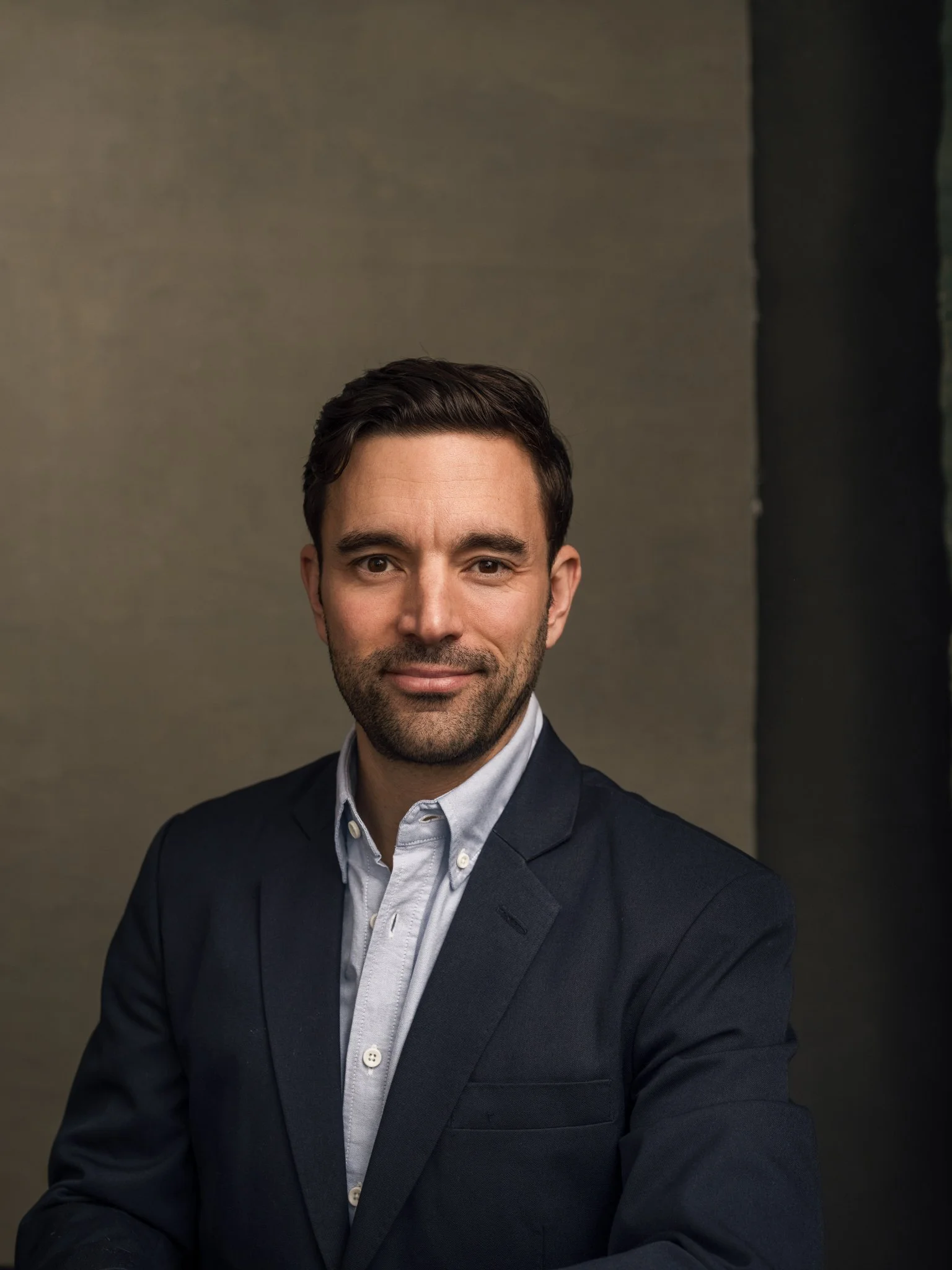 Professional portrait of a man with dark hair and beard, wearing a navy blazer and white shirt, standing against a plain background.