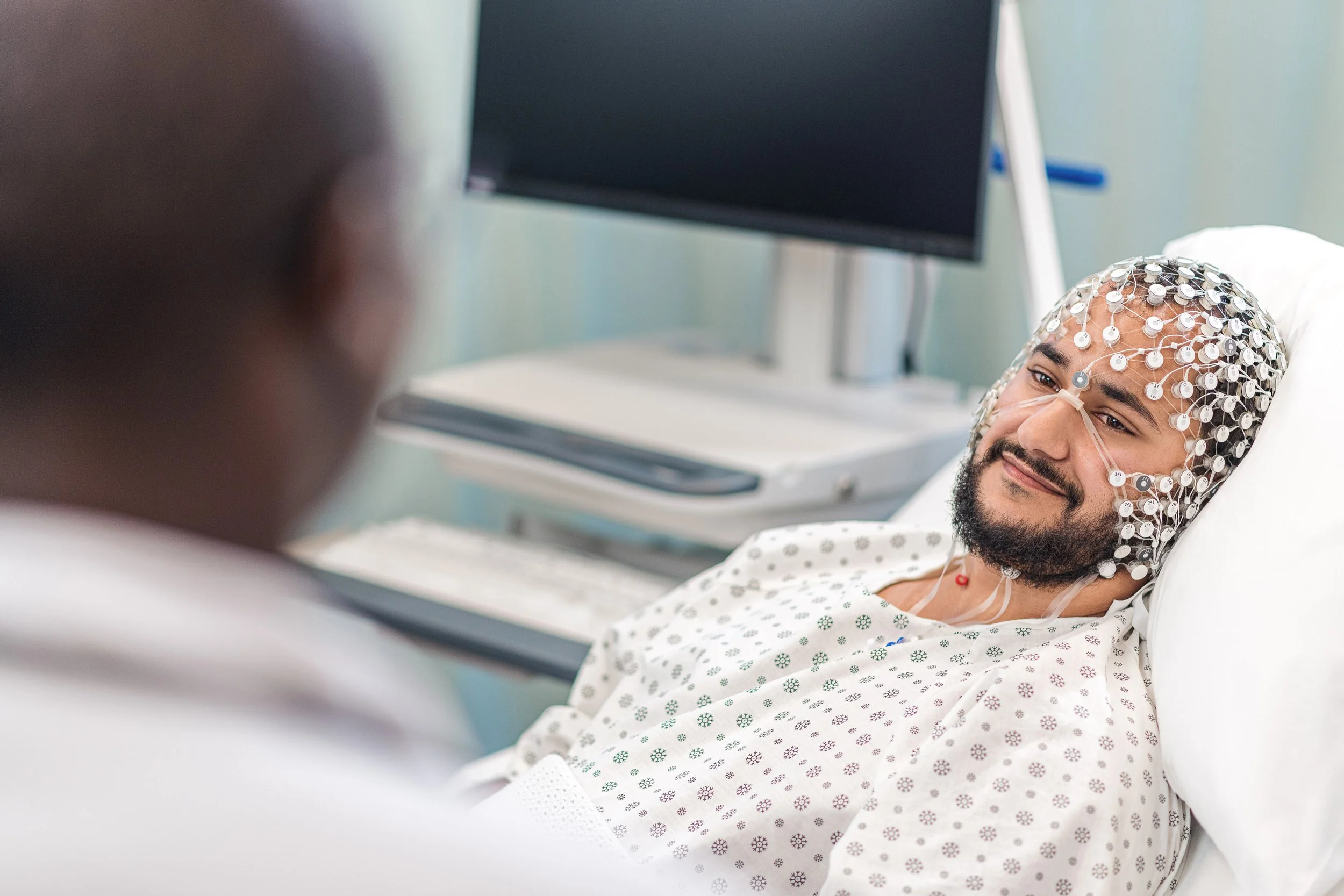 Male patient with a beard lying on a hospital bed, wearing a neurological monitoring cap with electrodes, smiling and speaking with a doctor in a medical facility.
