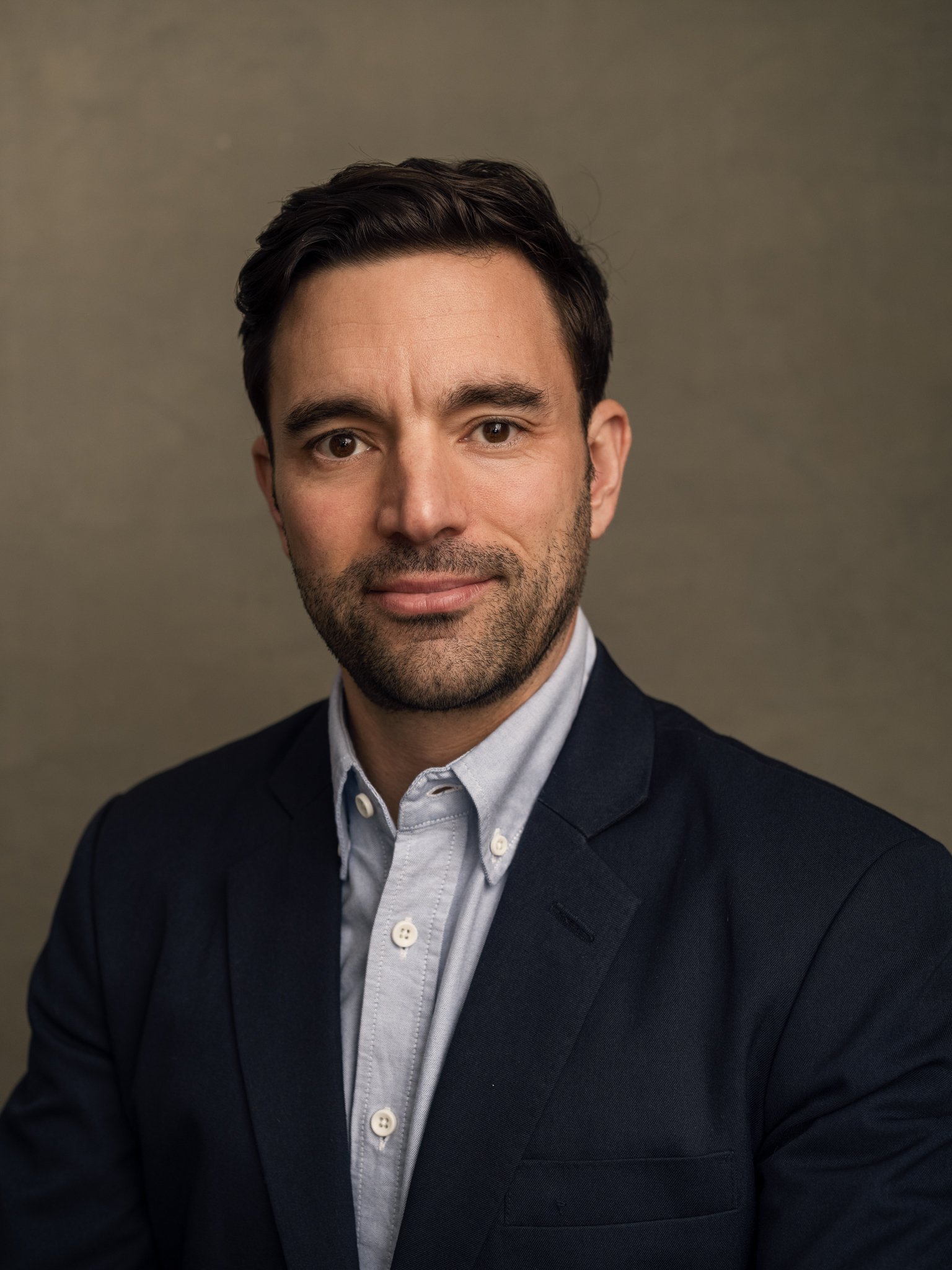 Professional headshot of a man with dark hair, beard, wearing a navy blazer and light-colored shirt, against a neutral background.