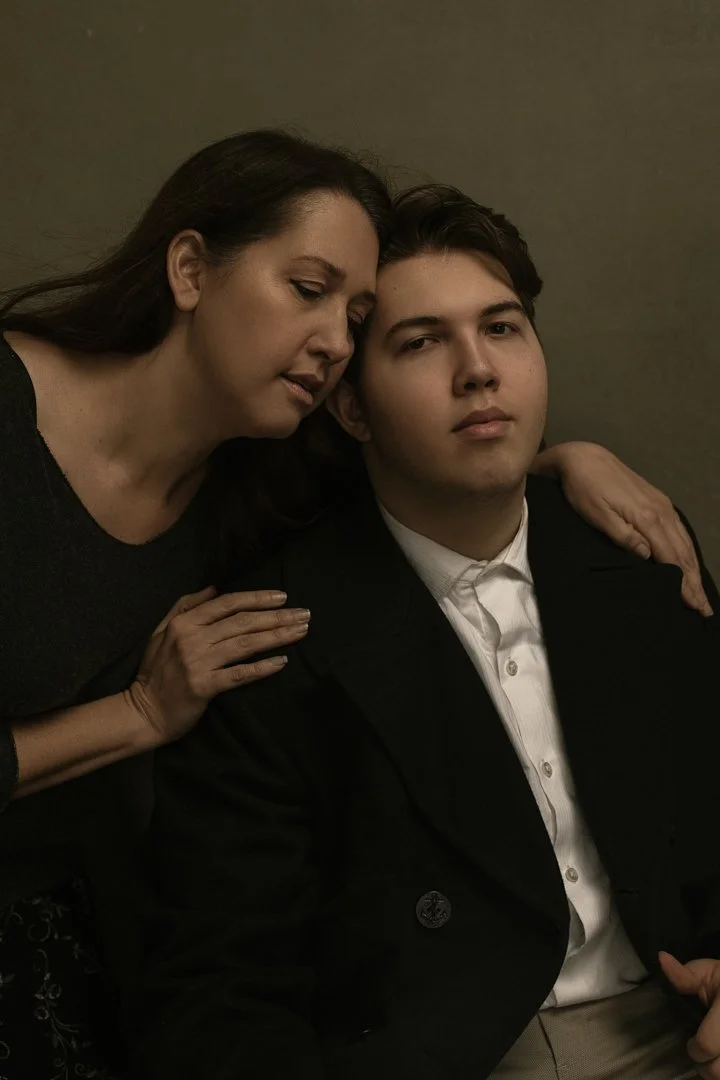 A woman and a young man pose closely together, with the woman resting her head on the young man's head and her hand on his shoulder, both looking at the camera in a dimly lit setting.