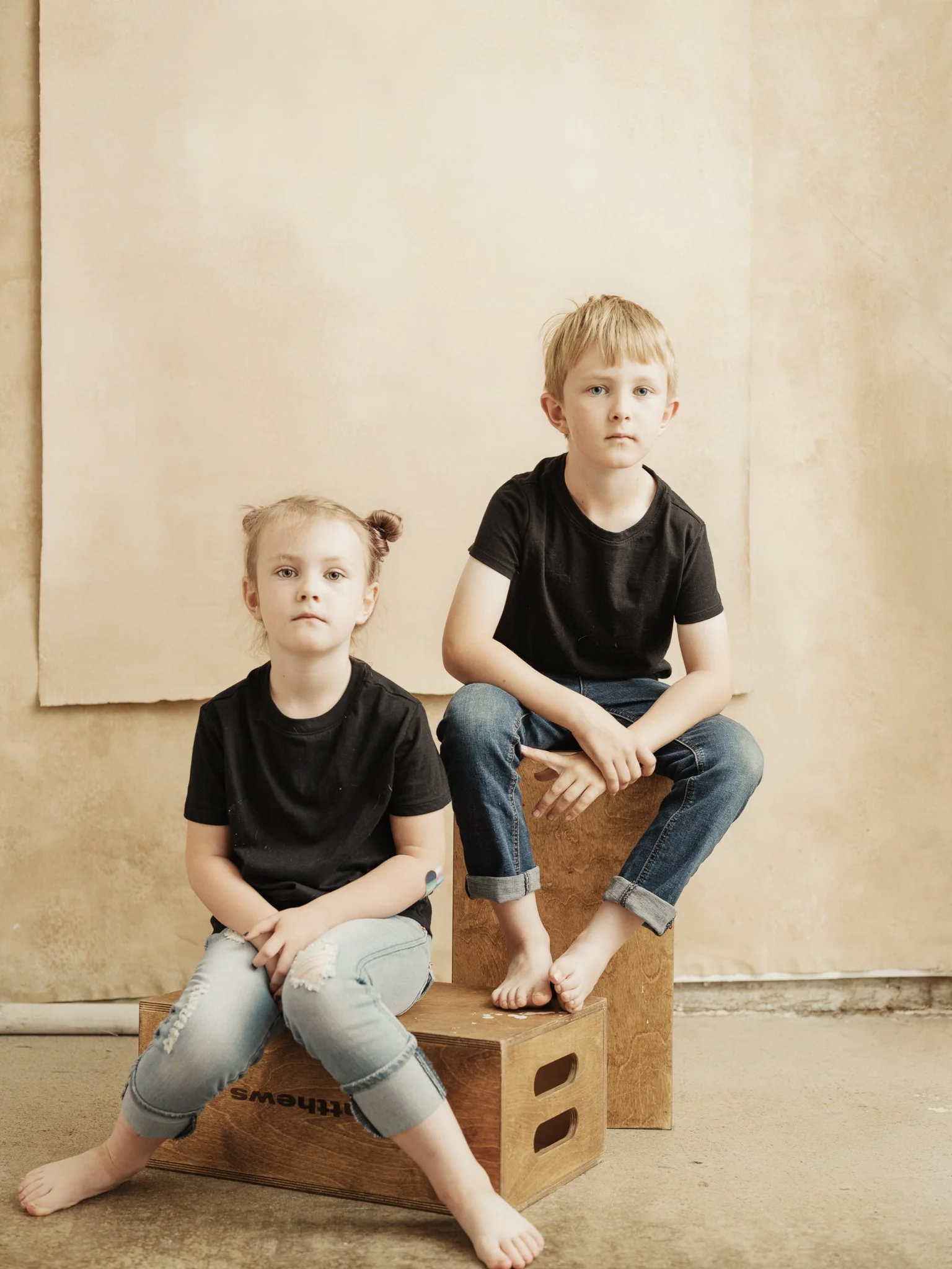 A boy and girl sitting on wooden boxes against a beige backdrop, both wearing black t-shirts and jeans, with the girl barefoot and sitting on the floor and the boy seated on a higher box, looking directly at the camera.