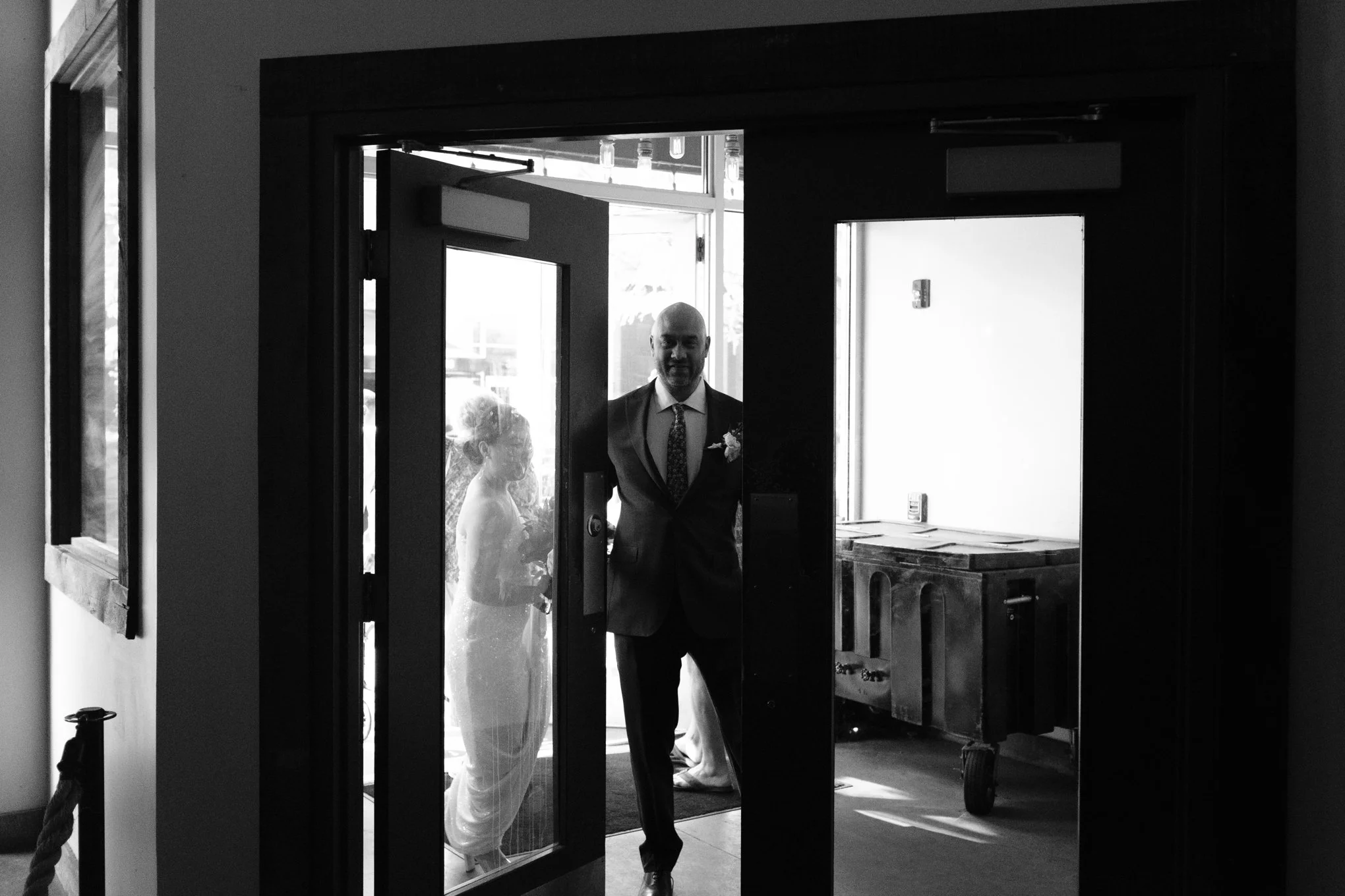 A black and white photo showing a man in a suit and a flower boutonniere standing at the entrance of a building, with a woman in a dress and a flower in her hair standing behind him, visible through a glass door.