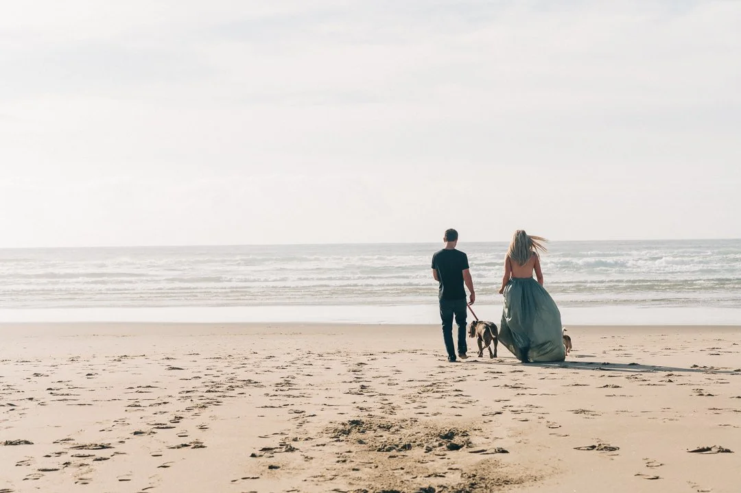 A couple walking with dogs on a sandy beach near the ocean, under a partly cloudy sky.