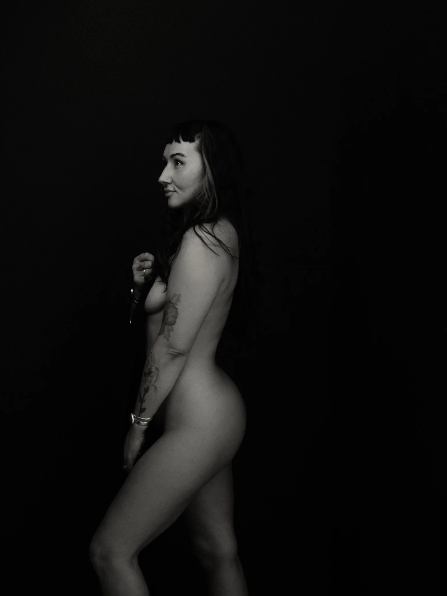 A black and white photo of a nude woman standing sideways against a dark background, with her gaze directed to the left. She has tattoos on her left arm and shoulder, dark hair with bangs, and is holding part of her hair with her left hand.