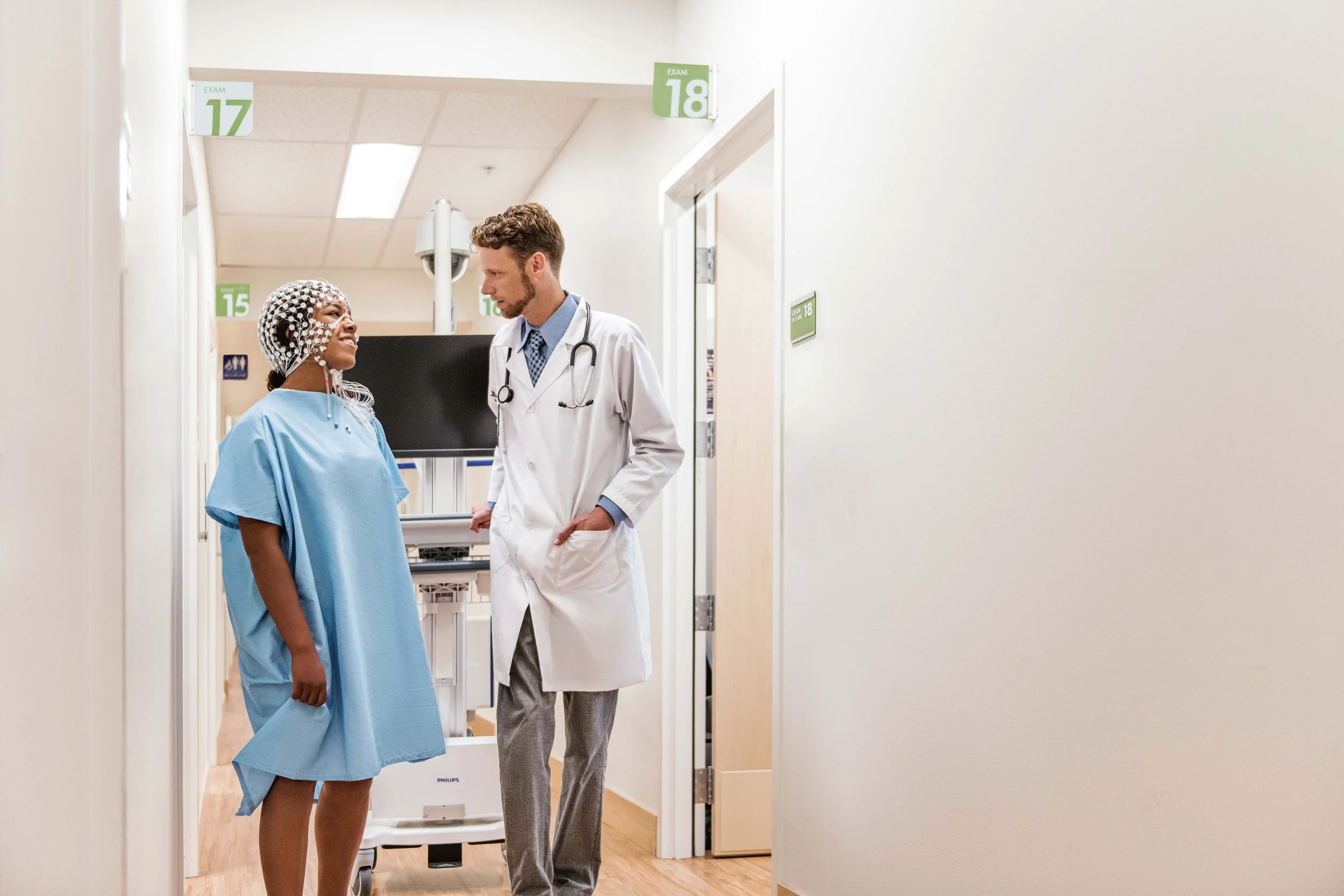 A female patient in a hospital gown and hairnet smiling and talking with a male doctor in a hospital hallway.