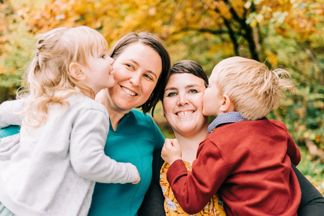Two women, one with dark hair and one with short hair, smiling while two children, a girl and a boy, kiss their cheeks in an outdoor setting during autumn.