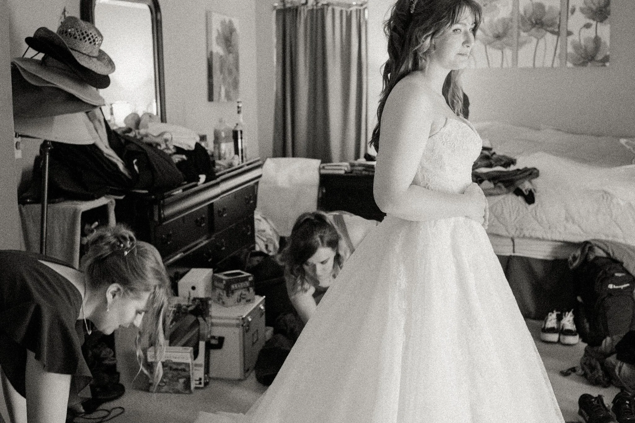 A woman in a wedding dress standing in a bedroom with two women kneeling and organizing items on the floor.