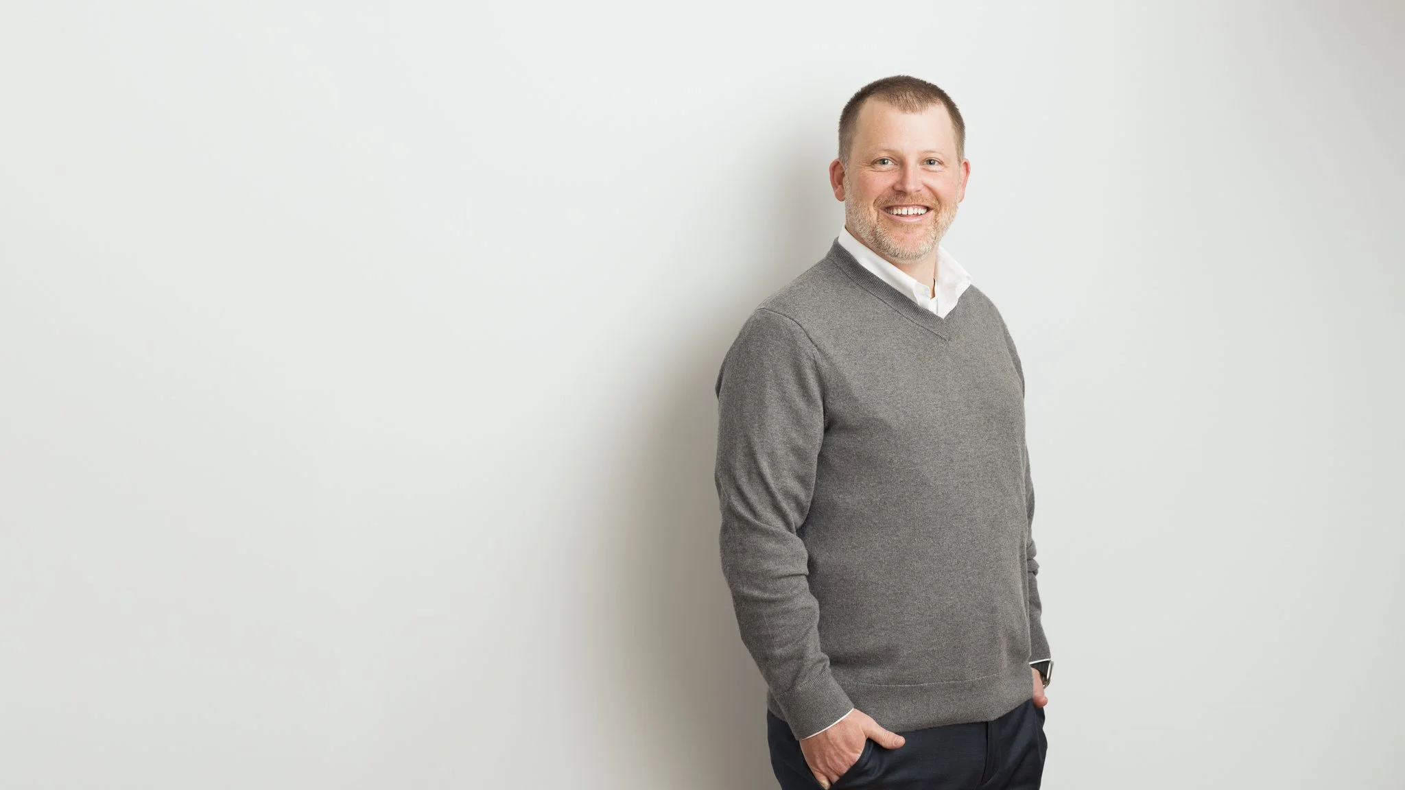 A smiling man with short hair and a beard, wearing a gray sweater over a white collared shirt, standing against a plain white background.