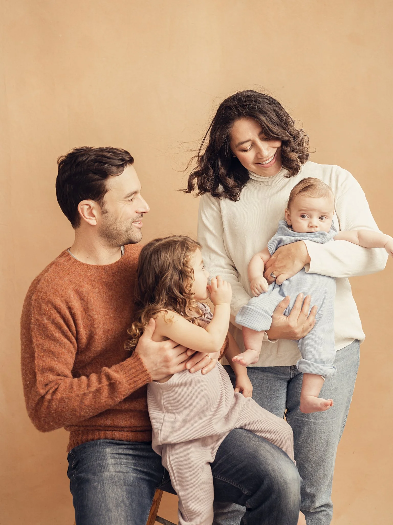 A family of four, including a father, mother, young daughter, and baby, with the mother holding the baby and the father holding the daughter, smiling and sitting against a neutral background.