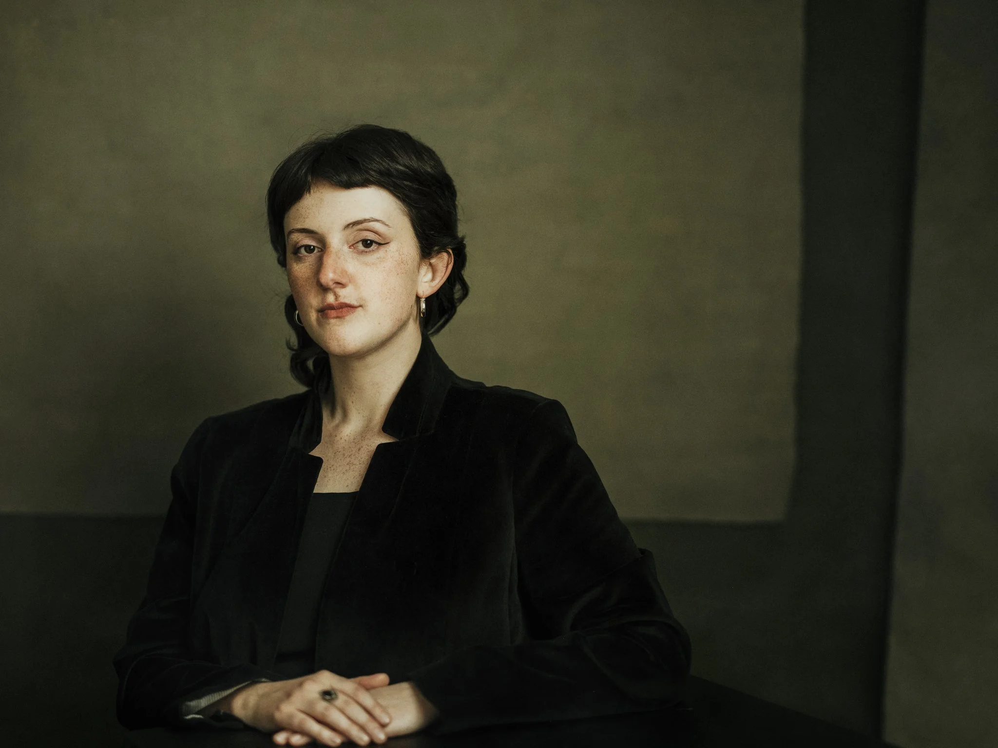 Professional headshot of a woman with short dark hair, wearing a black blazer with a lapel collar, sitting at a table with her hands folded. She is looking at the camera with a neutral expression, in a dimly lit room with a dark, textured background.