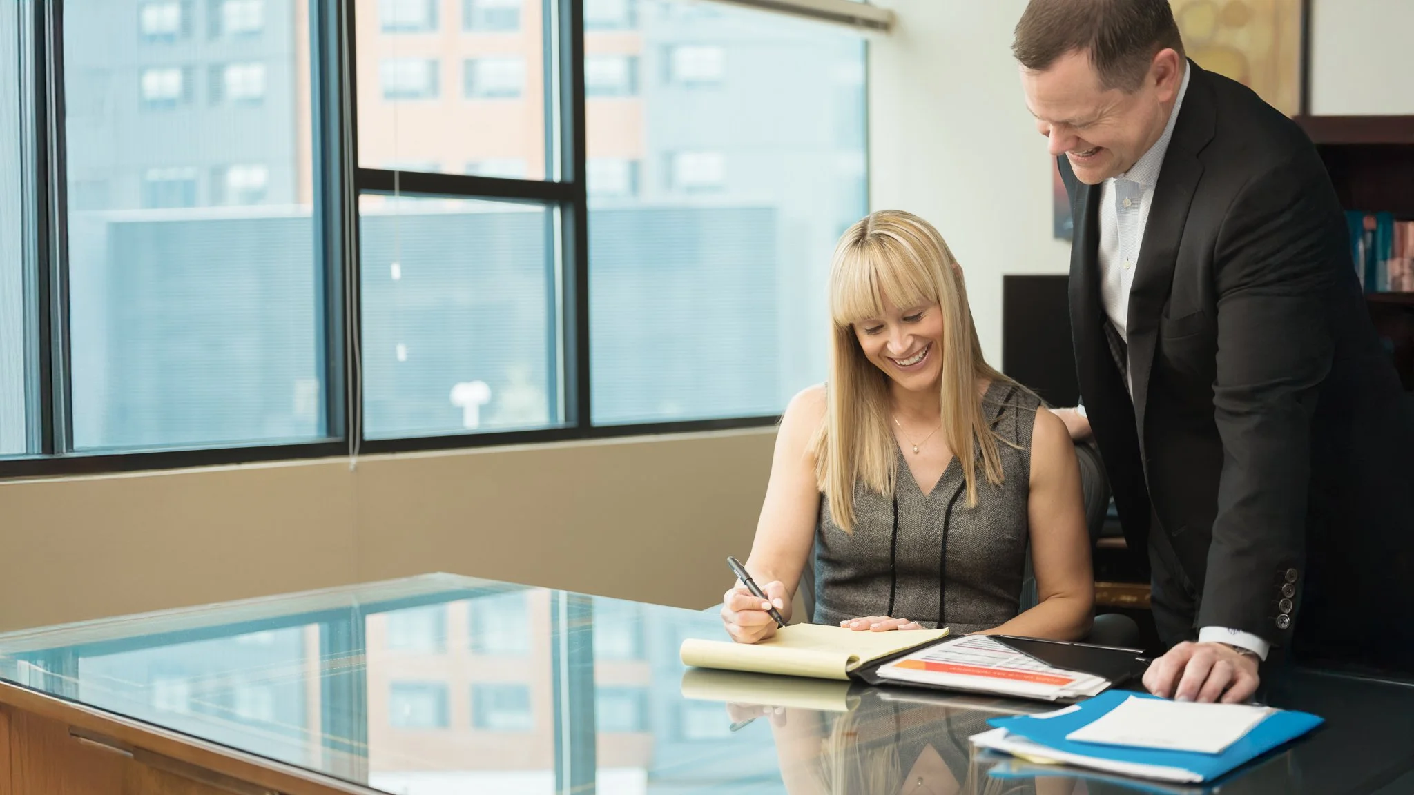 A woman sitting at a desk, signing a document, smiling while a man in a suit leans over her, laughing. The scene is inside an office with large windows and city buildings outside.