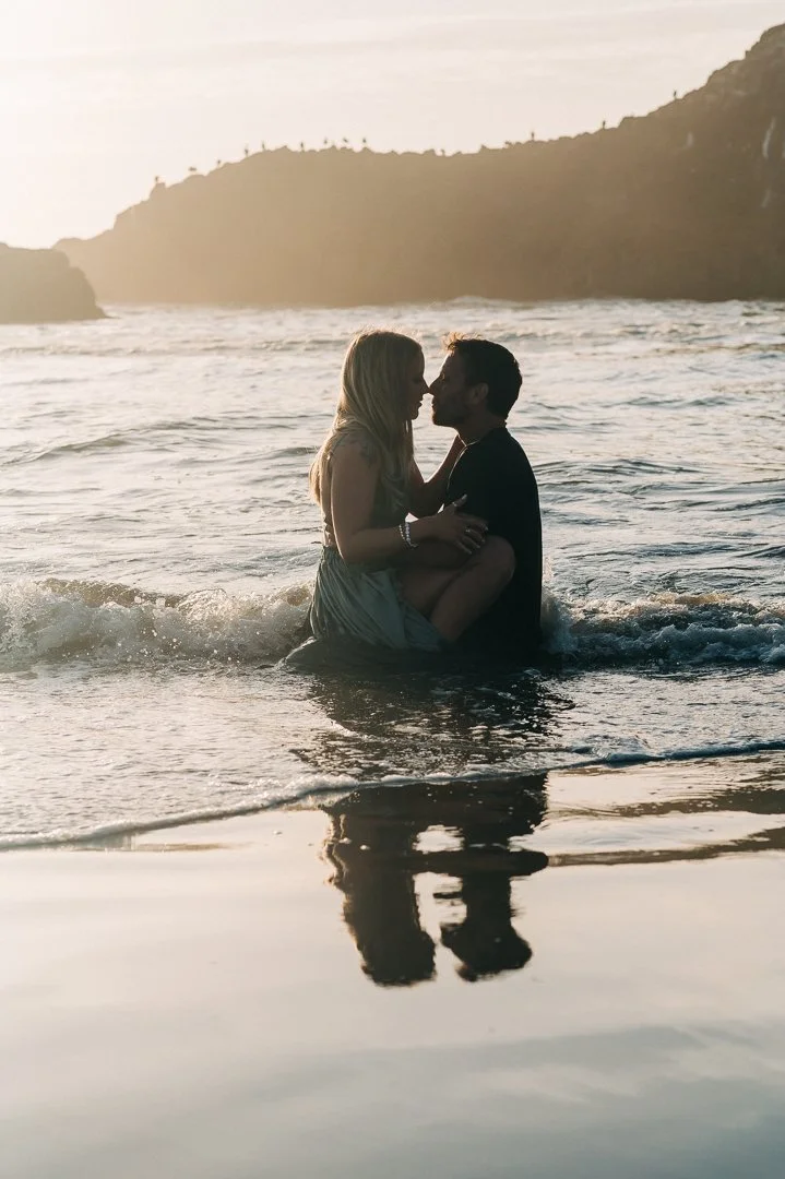 A couple sitting in the water at the beach, about to kiss, with a sunset and hillside in the background.