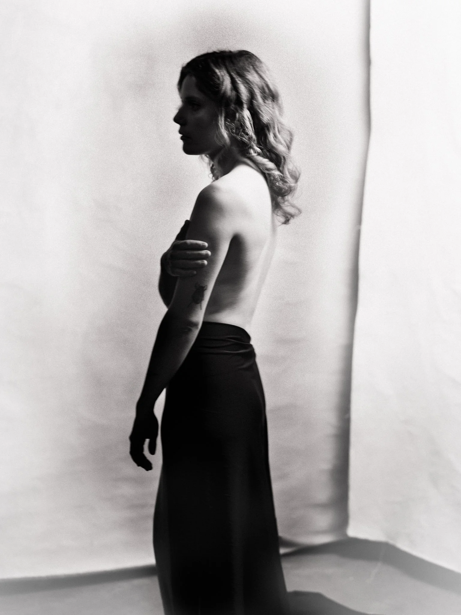Black and white photo of a woman with wavy hair, topless, wearing a long, dark skirt, standing sideways with her arms crossed over her chest, against a plain background.