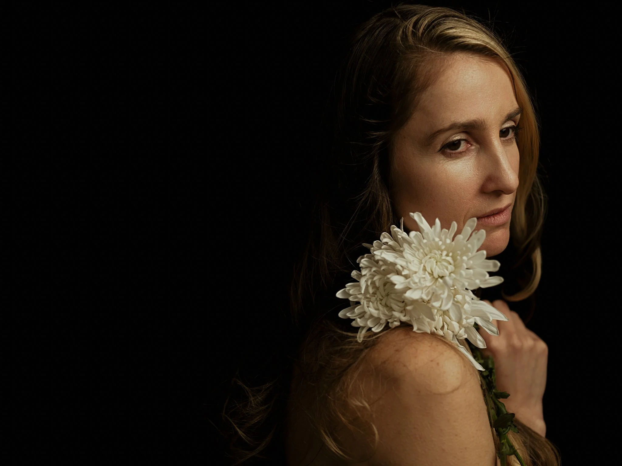 A woman with long, wavy hair holding white flowers on her shoulder against a black background.