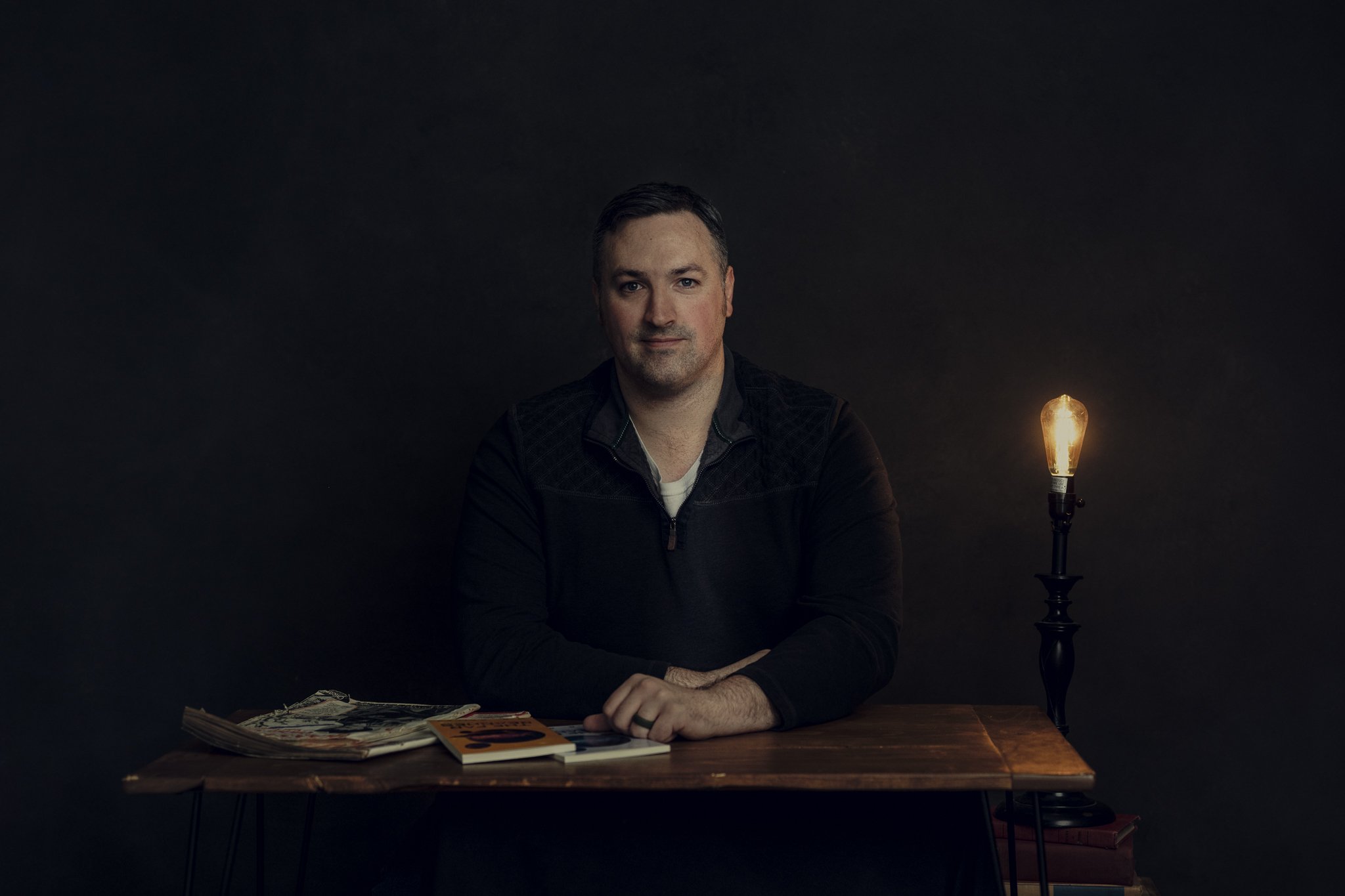 A man sitting at a wooden table in a dimly lit room, with magazines and books on the table, illuminated by a vintage-style table lamp with an exposed filament bulb.